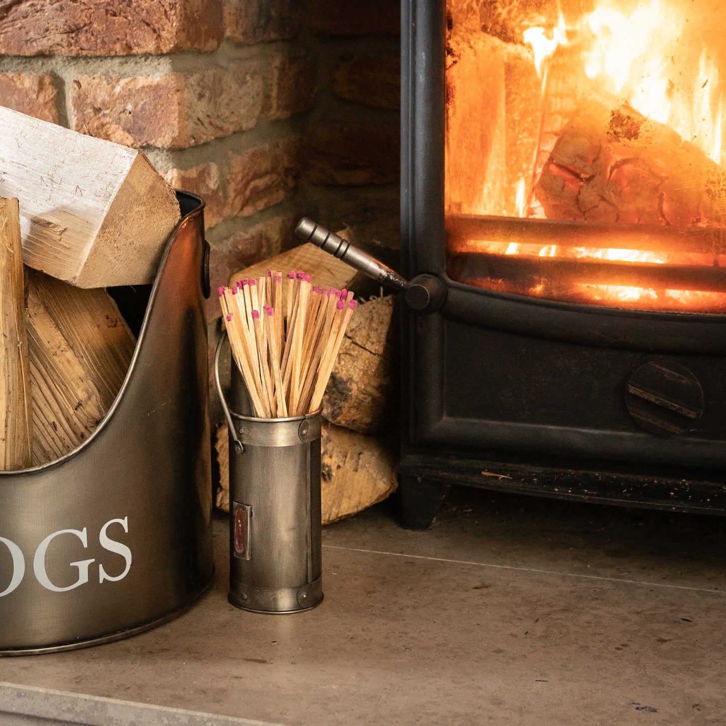 Antique pewter match holder with pink-tipped matches, elegantly placed next to a glowing fireplace and wooden logs.