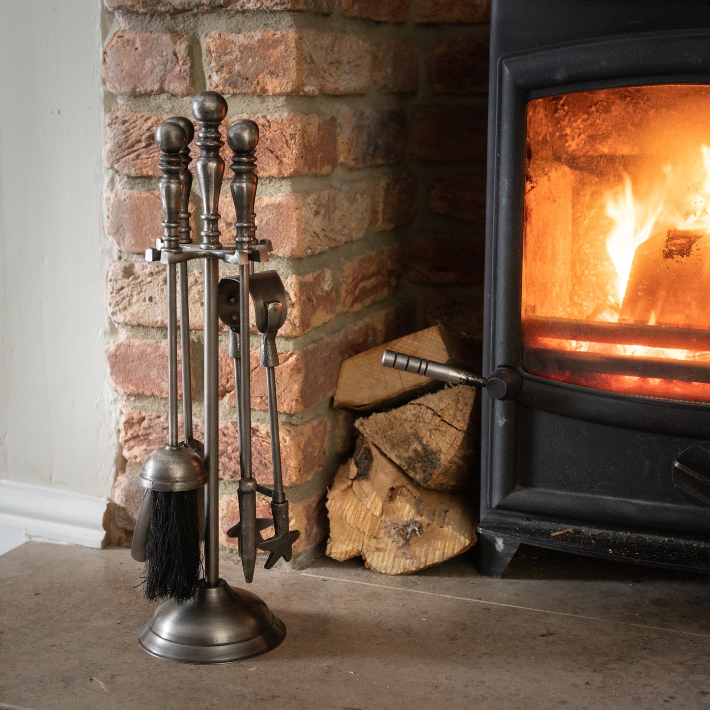 Antique pewter companion set stands beside a glowing fireplace, featuring elegant tools with ornate handles and a chic stand.