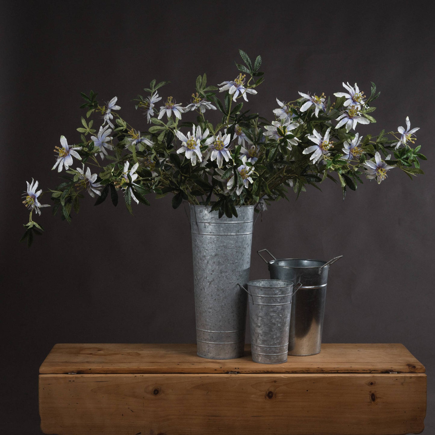 Lifelike Passion Flower Spray features elegant white petals and lush green foliage, displayed in a rustic metal vase.