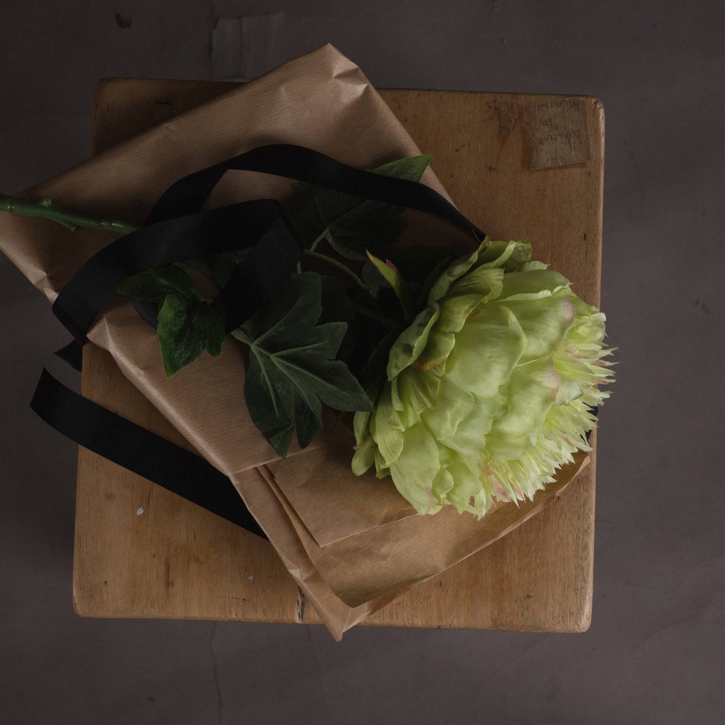 Sage-green peony flower with detailed petals rests on wrapped paper, accented by black ribbons on a wooden surface.