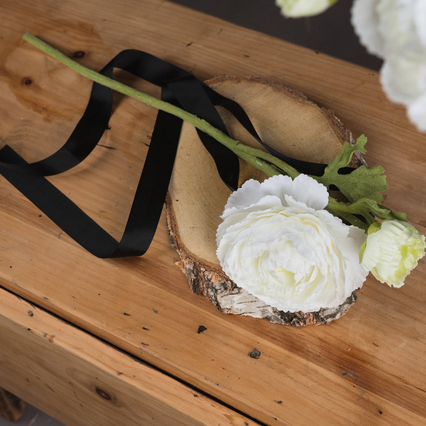 Elegant white Ranunculus spray with detailed petals and a natural green stem, resting on a wooden slice with black ribbon accents.