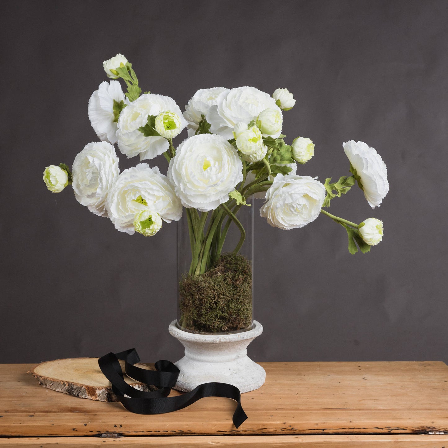 Elegant white ranunculus flowers in a glass vase with moss, complemented by a black ribbon, set on a wooden surface.