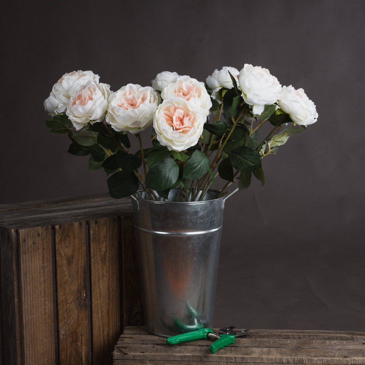 A silver bucket holds elegant artificial blush garden roses, featuring soft creamy petals and lush green leaves, against a dark backdrop.