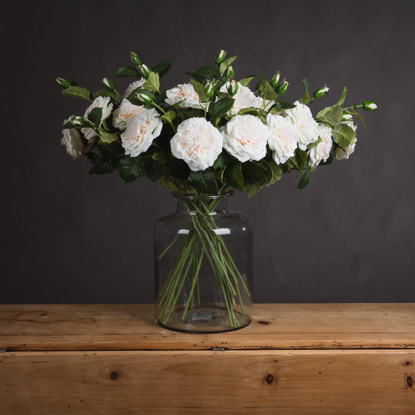 Luxurious artificial white garden roses in a glass vase, showcasing delicate blooms and lush greenery against a dark background.