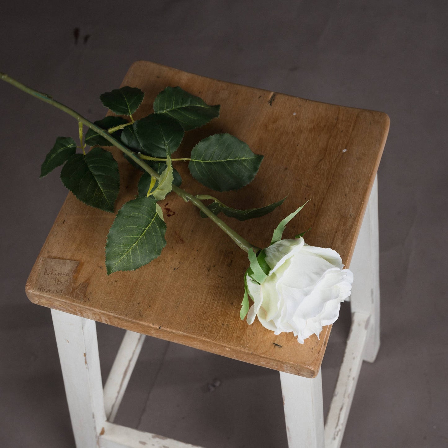 Elegant artificial white rose with realistic detailing, resting on a wooden stool, complemented by lush green leaves.