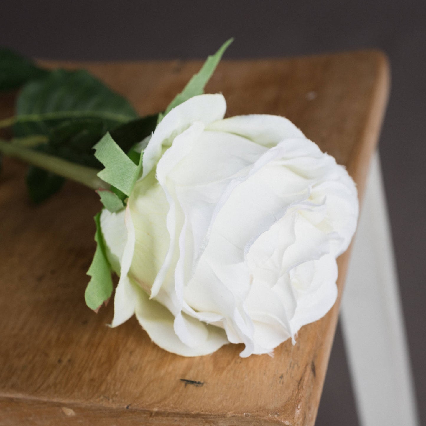 Realistic artificial white rose with detailed petals and leaves, resting on a wooden surface, enhancing elegant decor.