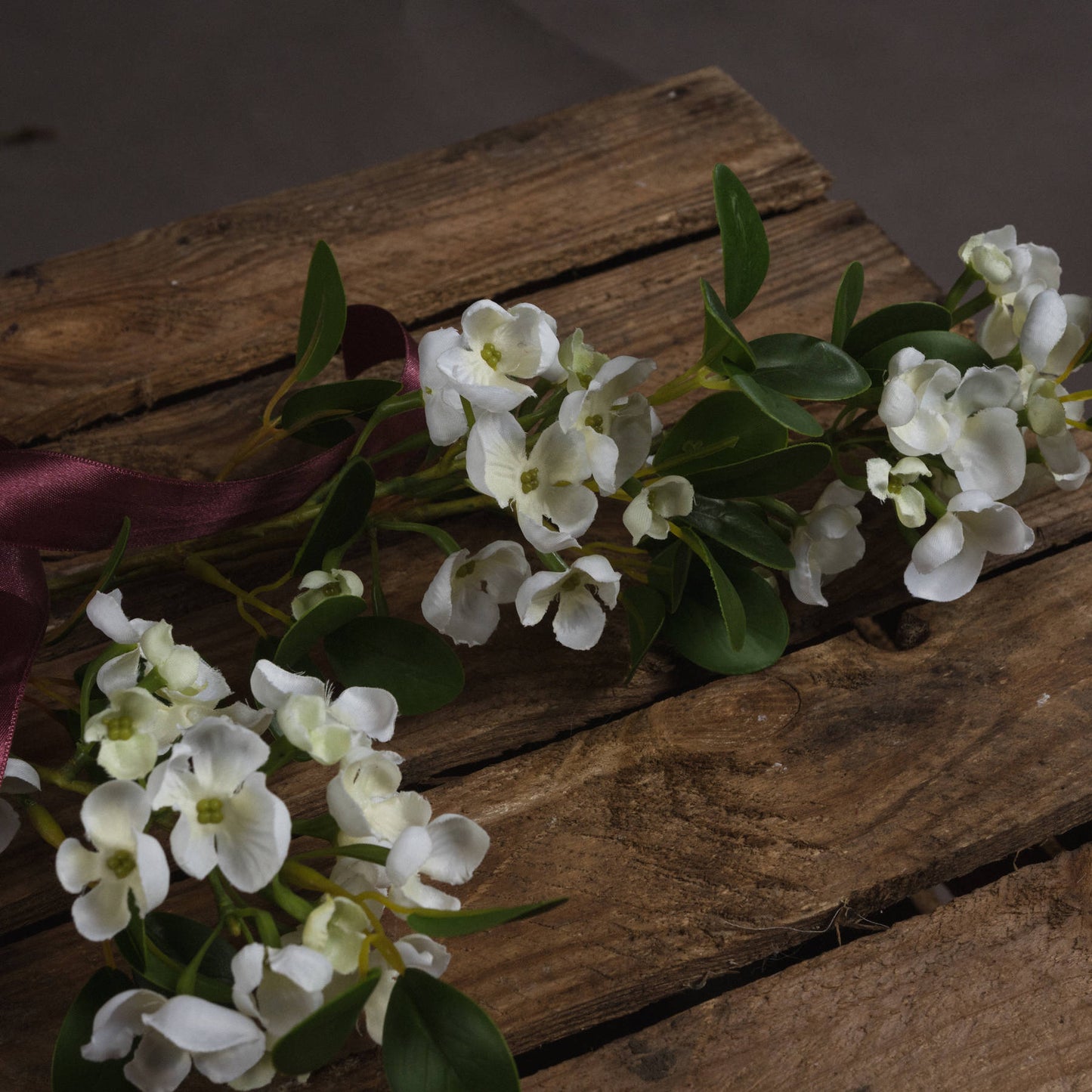 Lifelike white mock orange flowers with detailed petals and leaves, accented by a rich burgundy ribbon, on a rustic wooden surface.