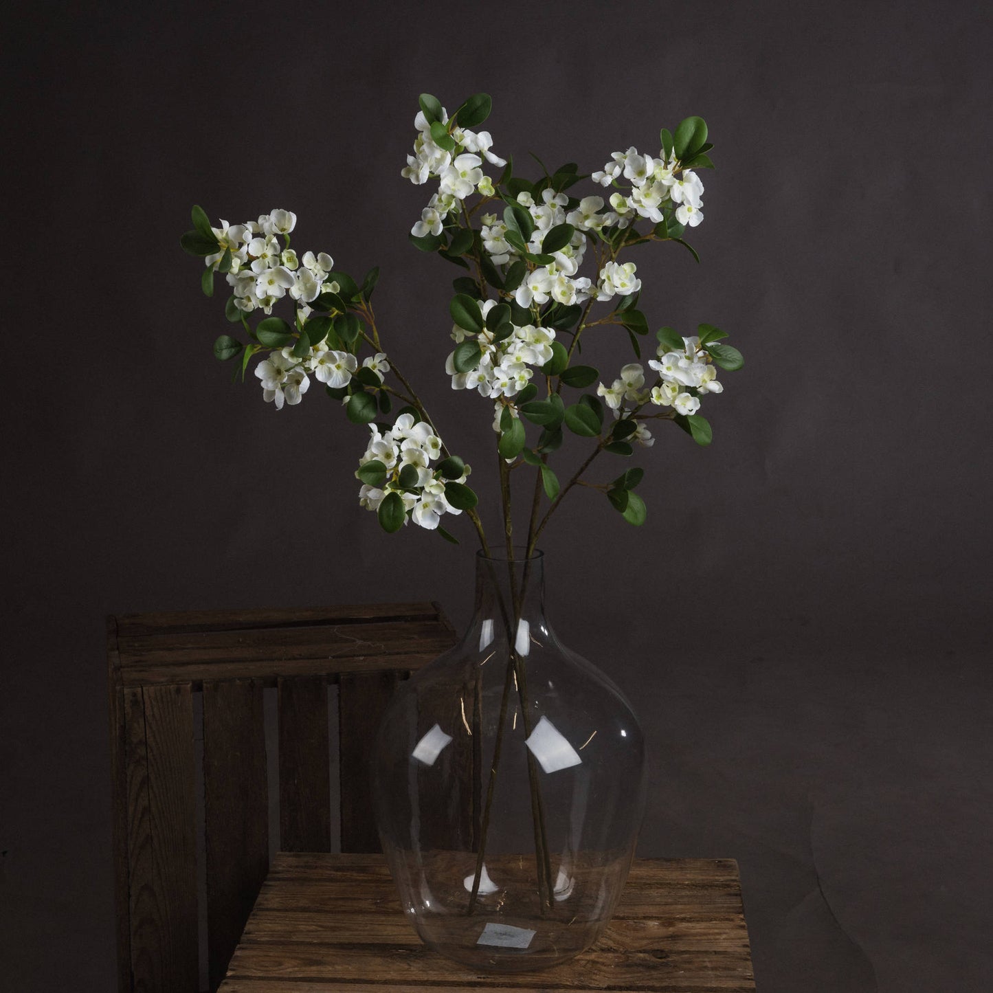 Elegant white mock orange flowers in a clear vase, set against a dark background, showcasing lush green leaves and lifelike detail.
