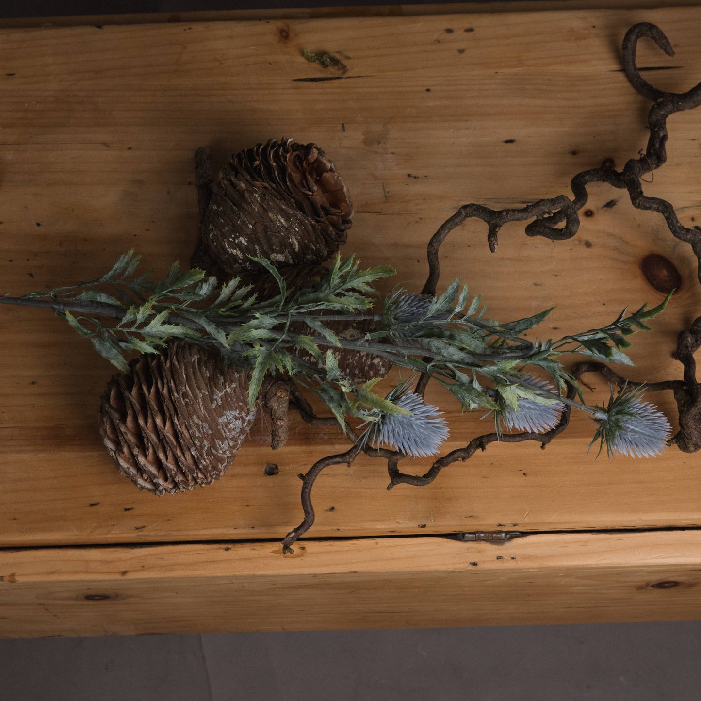 Lifelike blue thistle with powder-blue petals and green foliage, arranged with pine cones and twigs on a wooden surface.