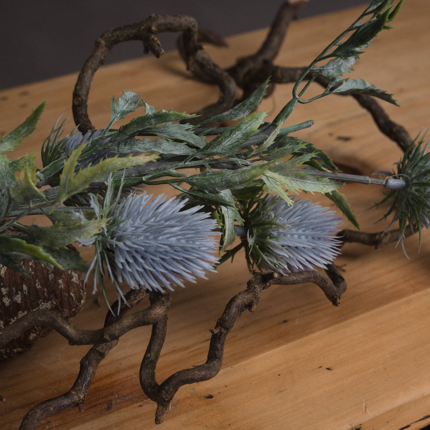 Lifelike blue thistle with textured powder-blue petals contrasts against green foliage and a rustic branch, enhancing decor.