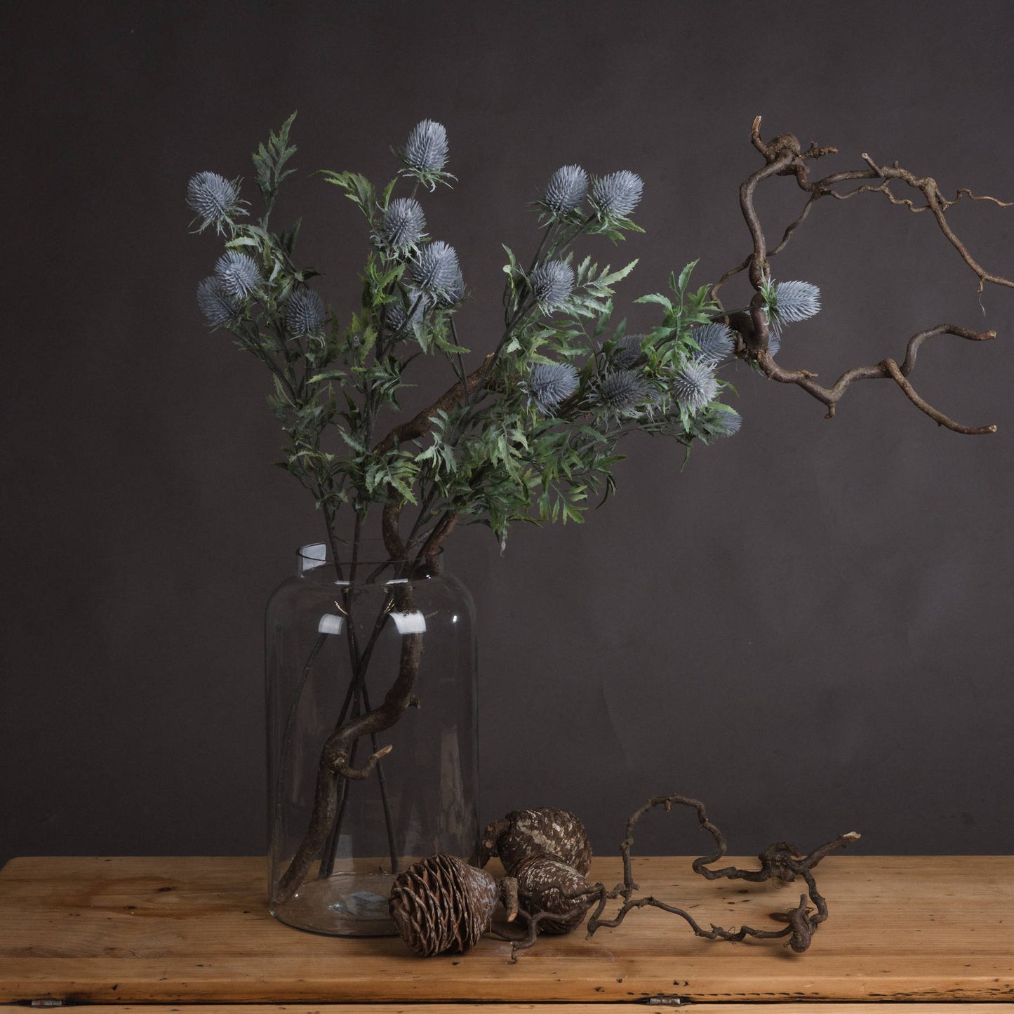 Lifelike blue thistle stems in a clear vase, surrounded by rustic accents on a wooden table, against a dark backdrop.