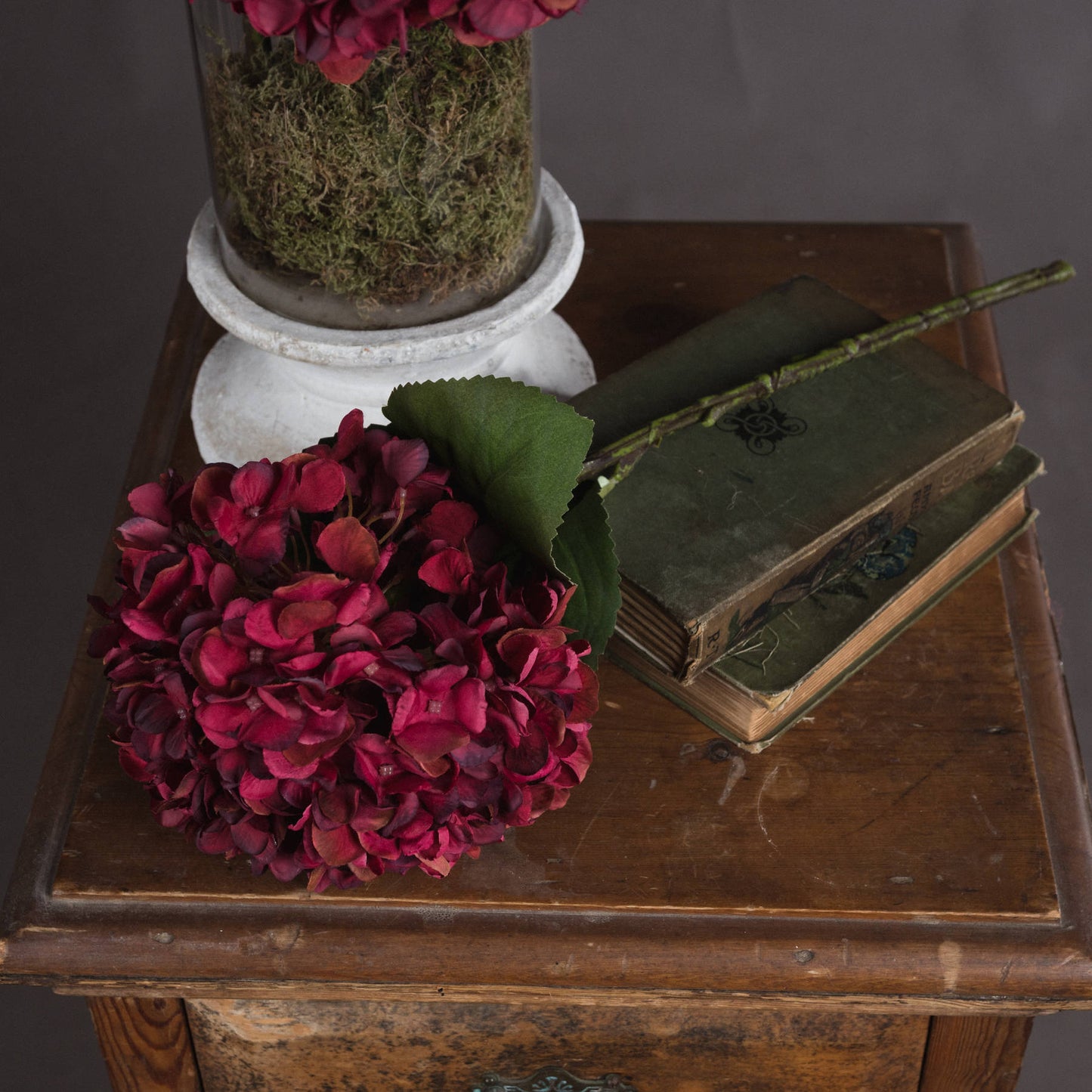 Rich ruby hydrangea bloom with hand-painted petals, nestled beside vintage books and a moss-filled glass vase, adding autumnal elegance.