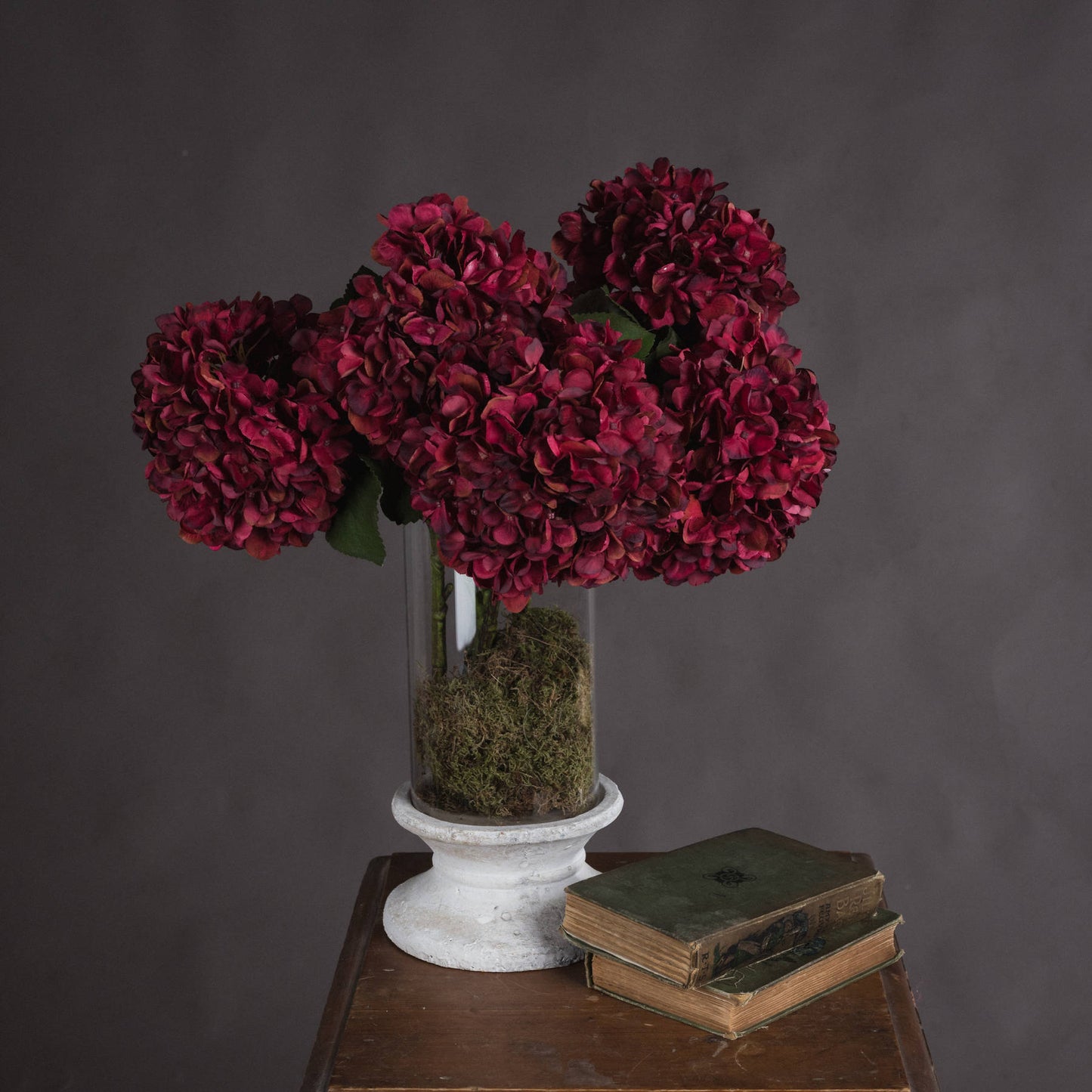 Deep ruby hydrangeas in a glass vase with moss base, set on a wooden table beside vintage books, creating an inviting autumn ambiance.