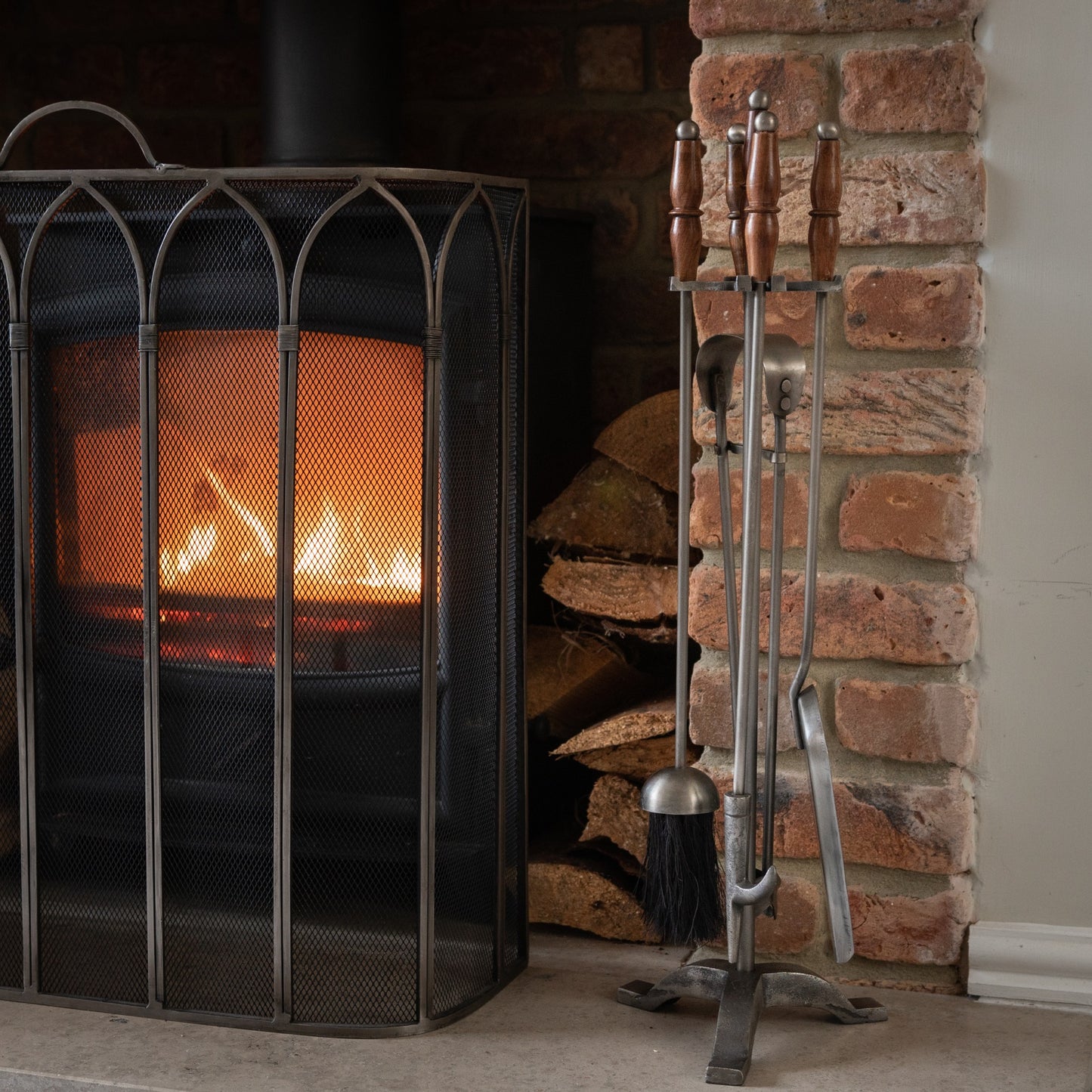 Hand-turned fire companion set in antique pewter with wooden handles, featuring a sophisticated design beside a glowing fireplace.