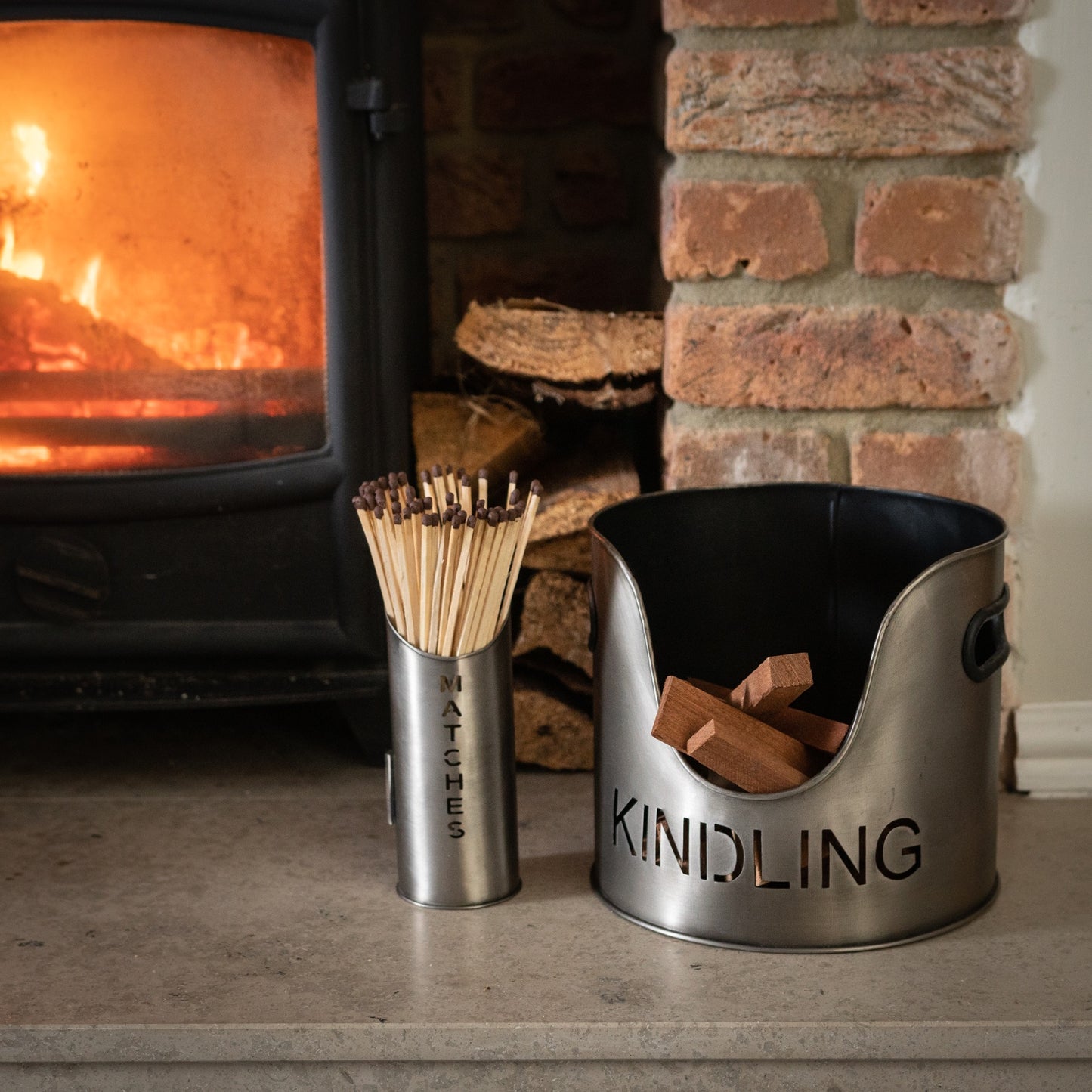Aged pewter finish kindling bucket with "KINDLING" cutout, alongside a matchstick holder labeled "MATCHES," both near a warm fireplace.
