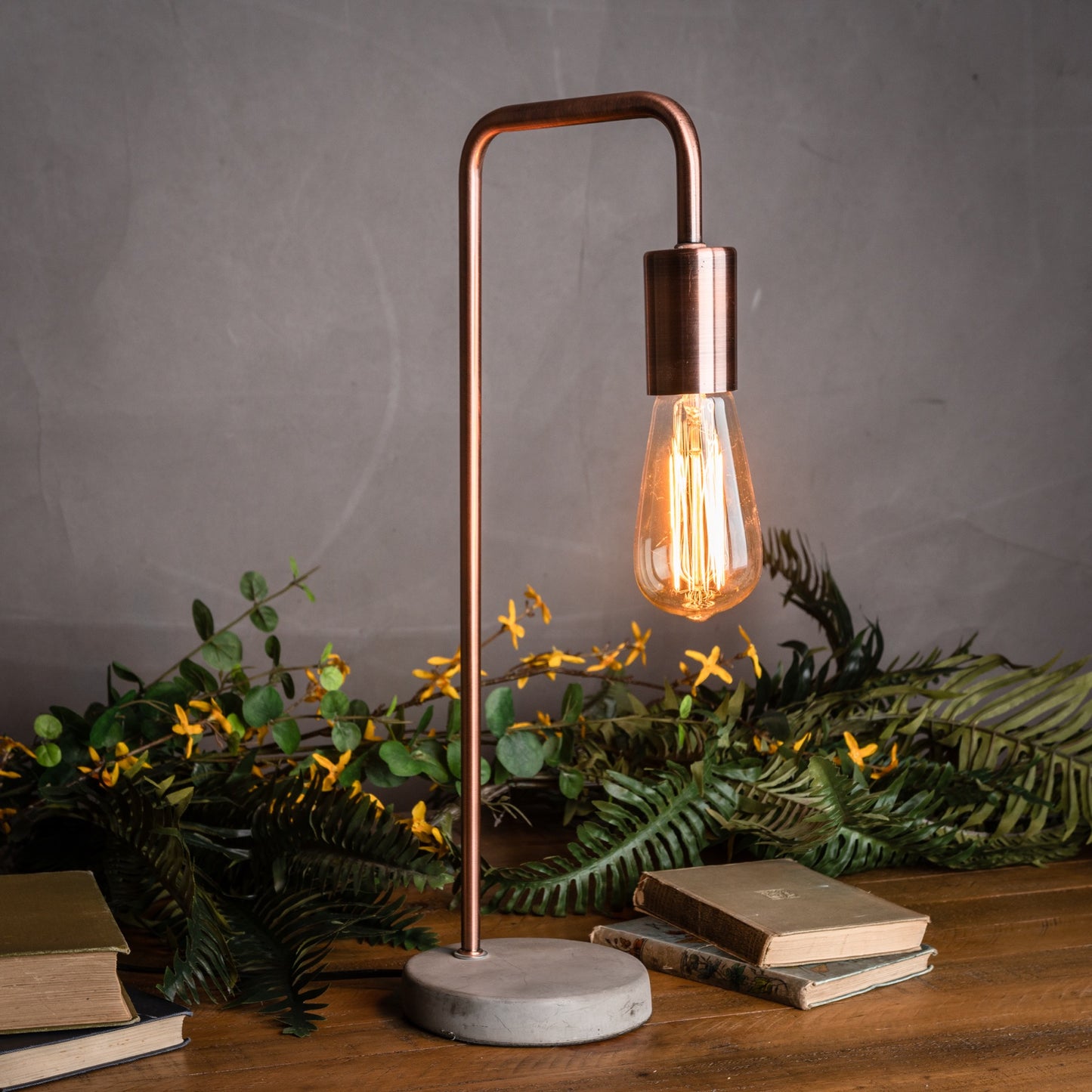 Copper industrial lamp with a stone base; sleek design, warm light, and surrounded by green foliage and books.