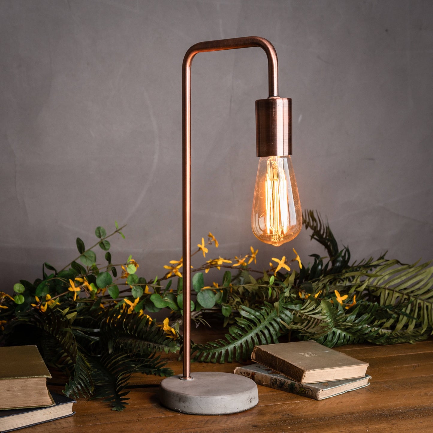 Copper industrial lamp with a stone base, featuring a warm light bulb and surrounded by greenery and vintage books.