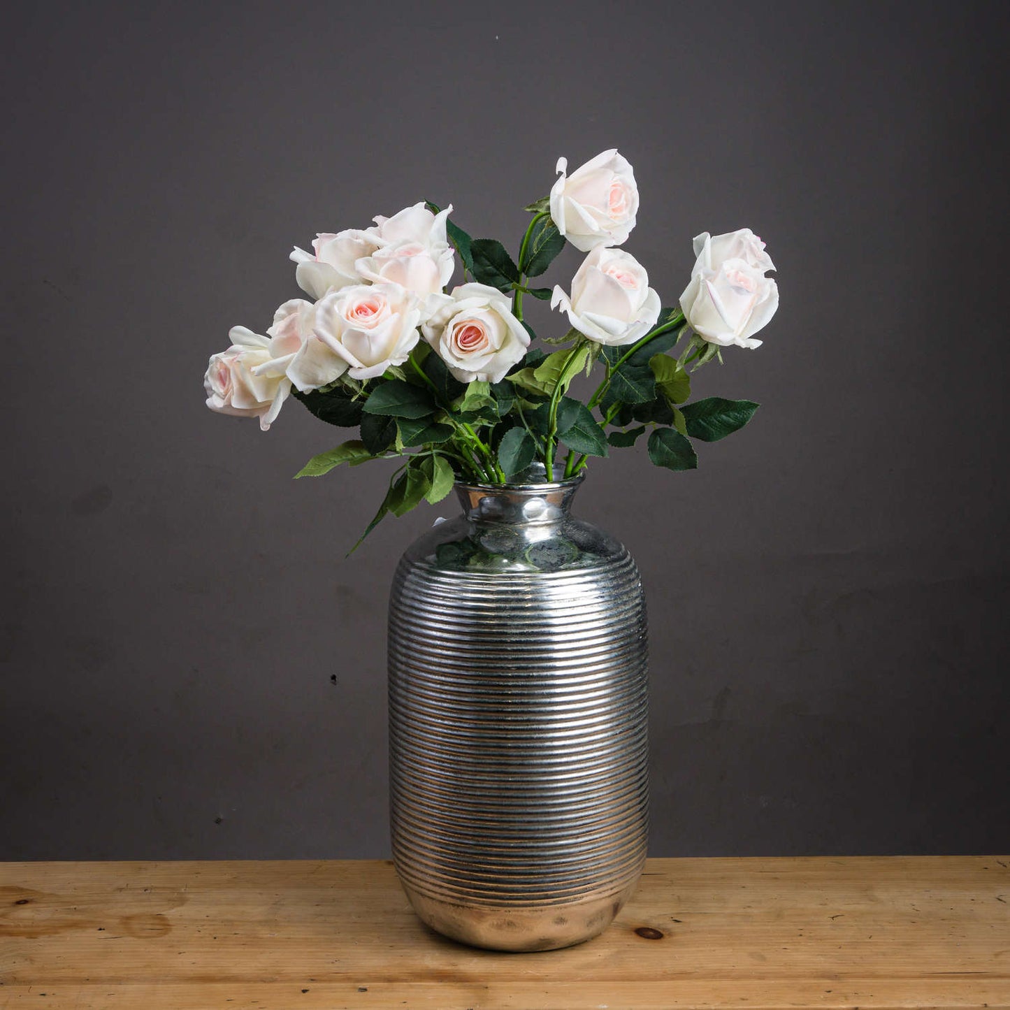 Soft blush pink artificial tea roses bloom gracefully among vibrant green leaves in a sleek, silver-striped vase.