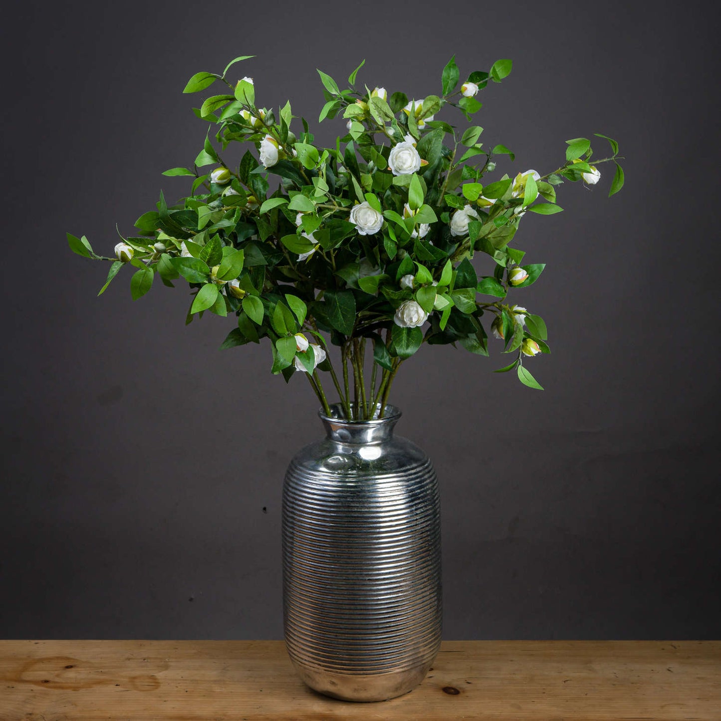 Cream gardenia stem with realistic cream petals and green leaves, arranged in a textured silver vase against a dark background.