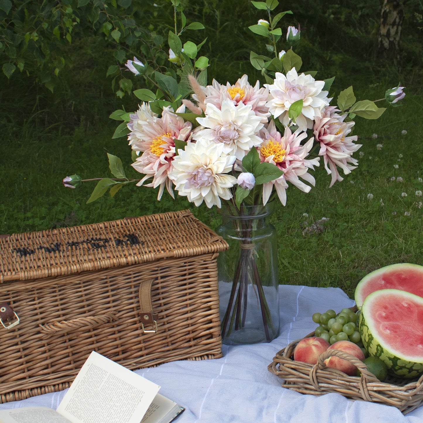 Lush pink artificial dahlia bouquet in a glass vase, surrounded by a woven basket, fruits, and an open book on a blanket.