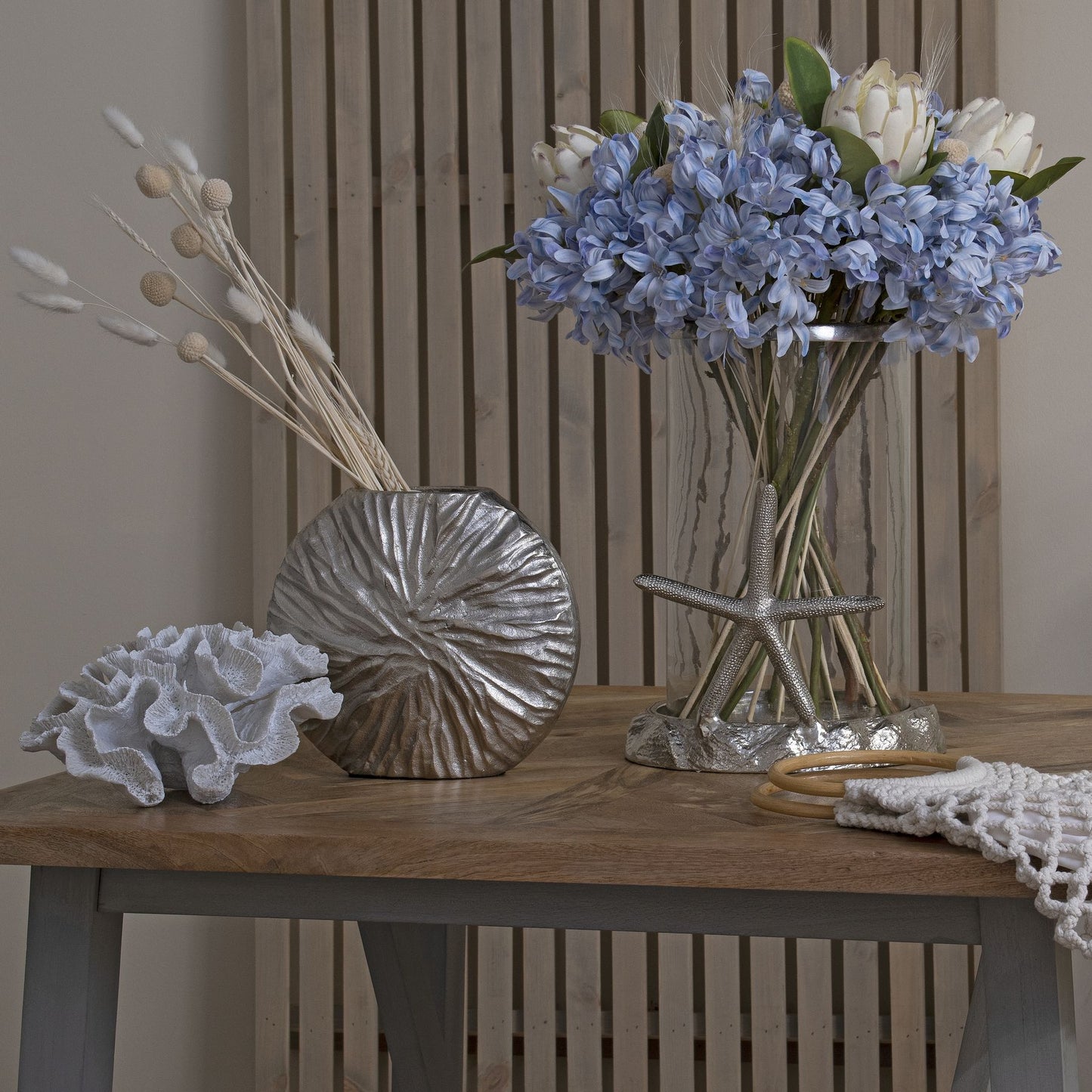 Light blue agapanthus flowers in a clear vase, complemented by silver decor and natural elements, creating a serene coastal ambiance.