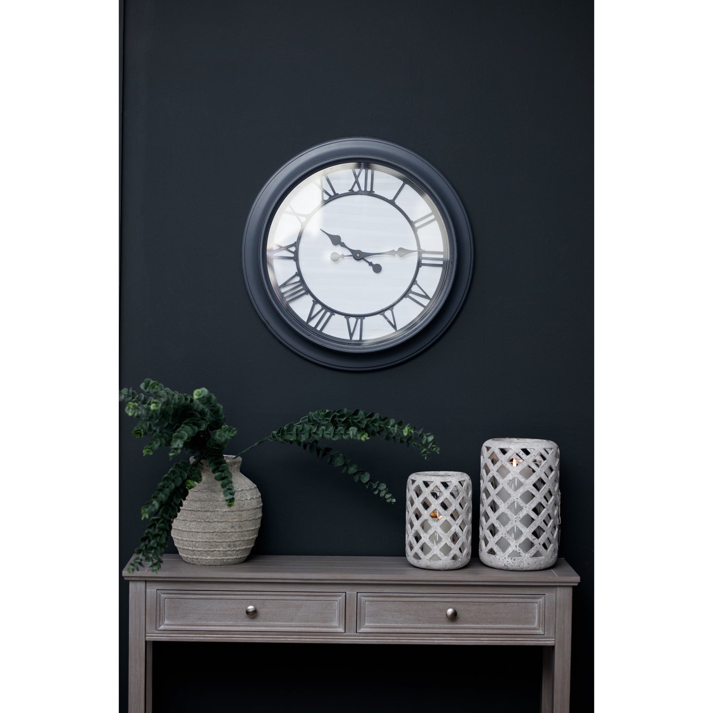 Sleek black-framed wall clock with Roman numerals, displayed above a light wooden console and decorative vases on a dark wall.