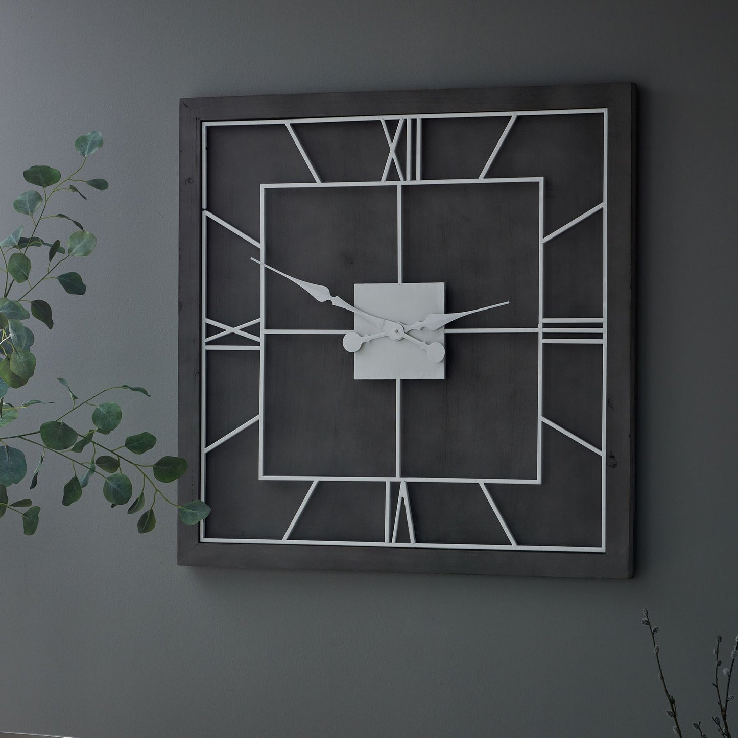 Square wall clock with a classic grey frame, white Roman numerals, and intricate design, mounted against a dark wall.