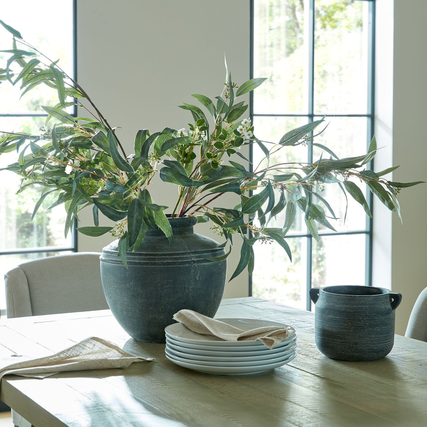 Realistic Seeded Eucalyptus Spray in a dark vase, featuring lush green leaves and seed pods, enhances a modern dining table setting.