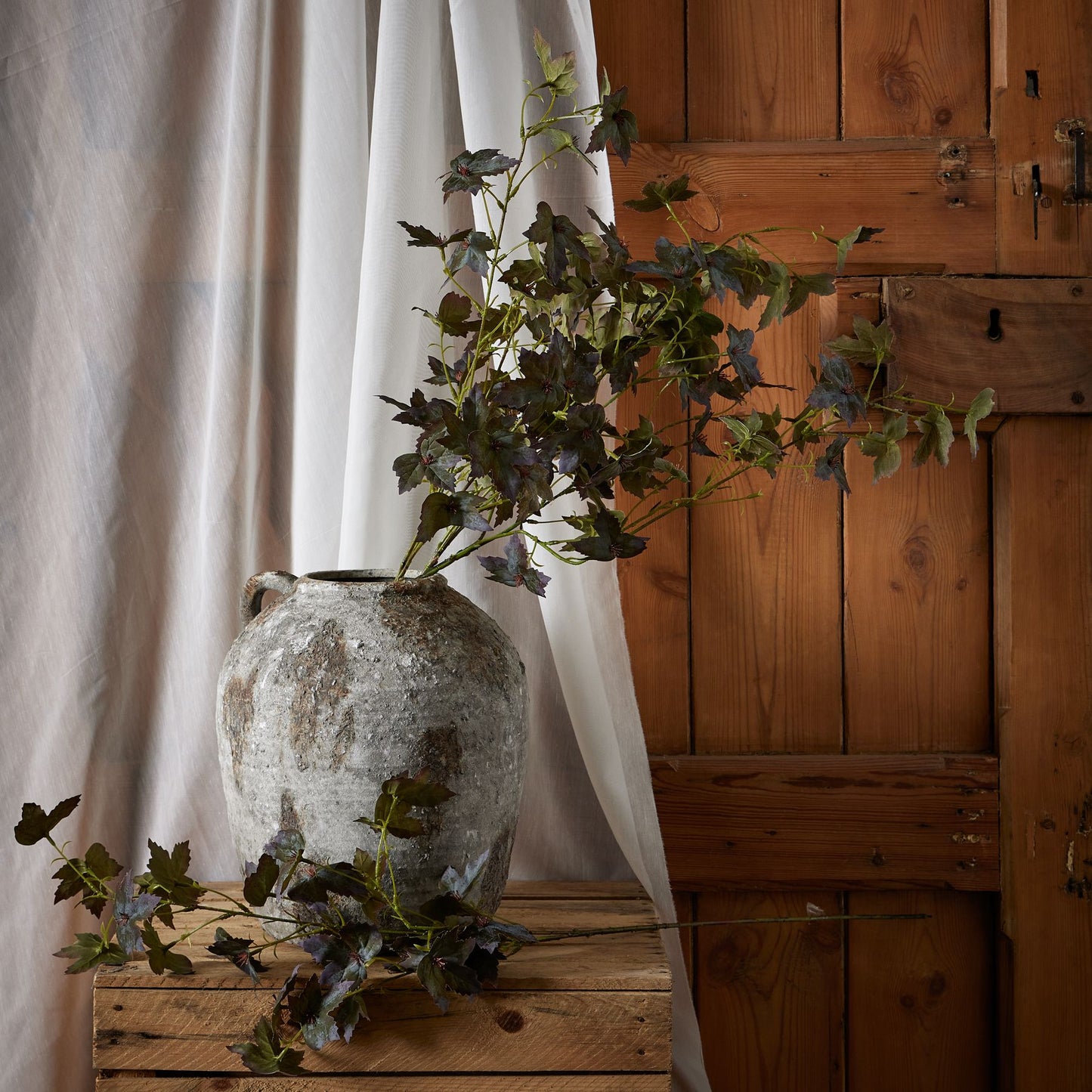 Variegated maple leaves in rich amber and burgundy hues extend from a rustic gray vase, set against a wooden backdrop.