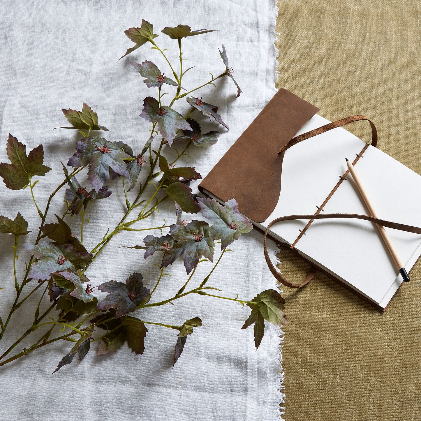 Variegated maple leaf stem featuring rich amber and deep burgundy hues, alongside a leather-bound notebook and pencil.