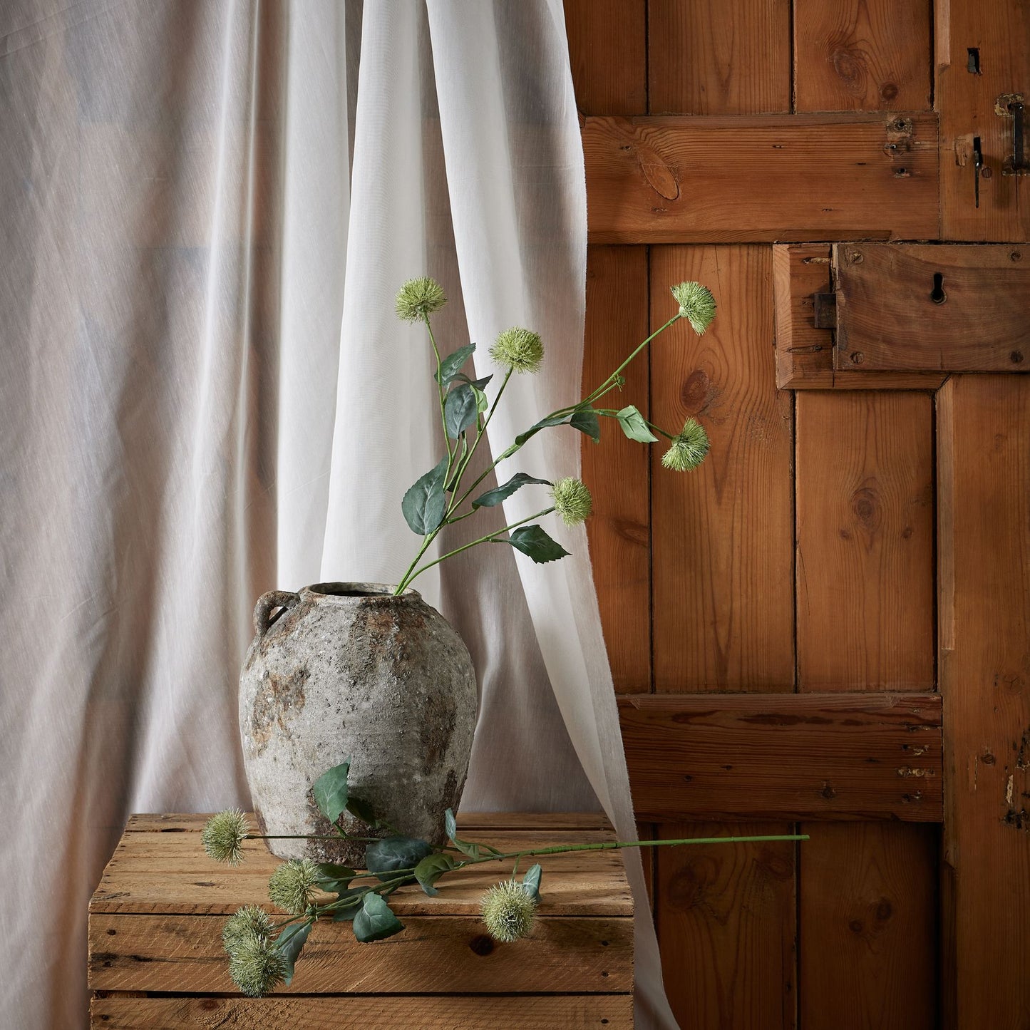 Green artificial thistle globe stem in a textured grey vase, set against light fabric and rustic wood background.