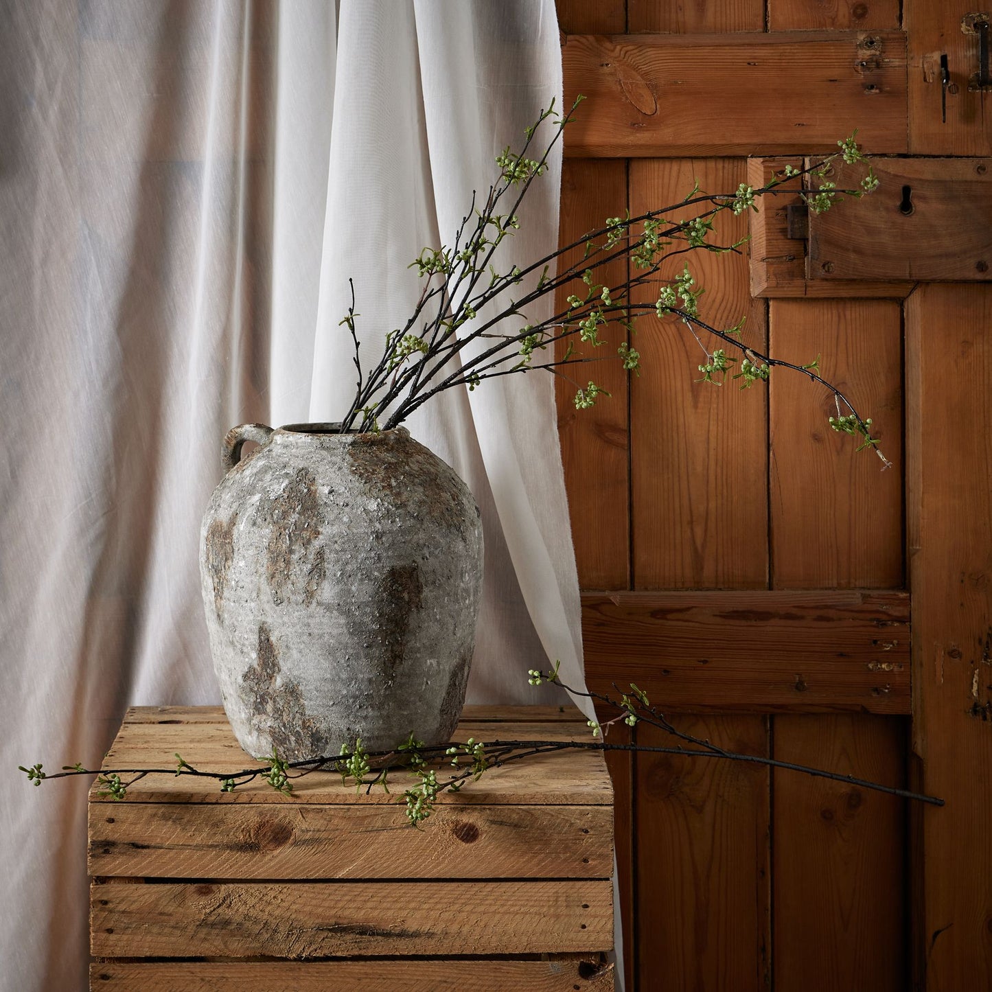 Green Berry Willow Stem features slender branches with vibrant green berries, arranged elegantly in a rustic gray vase on a wooden crate.