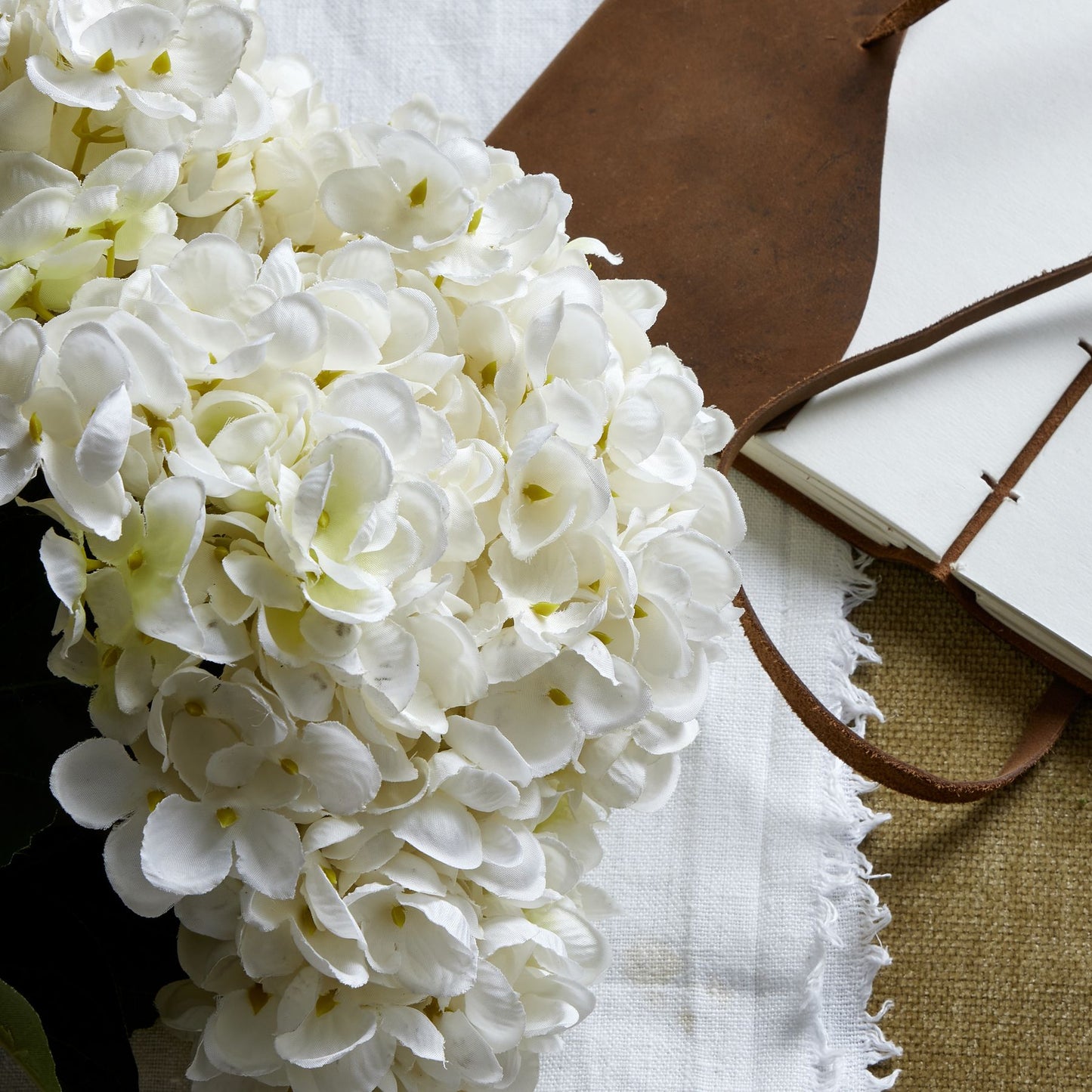 Pristine white hydrangea bunch with intricately crafted petals, laid on a textured background with a leather notebook and fabric.