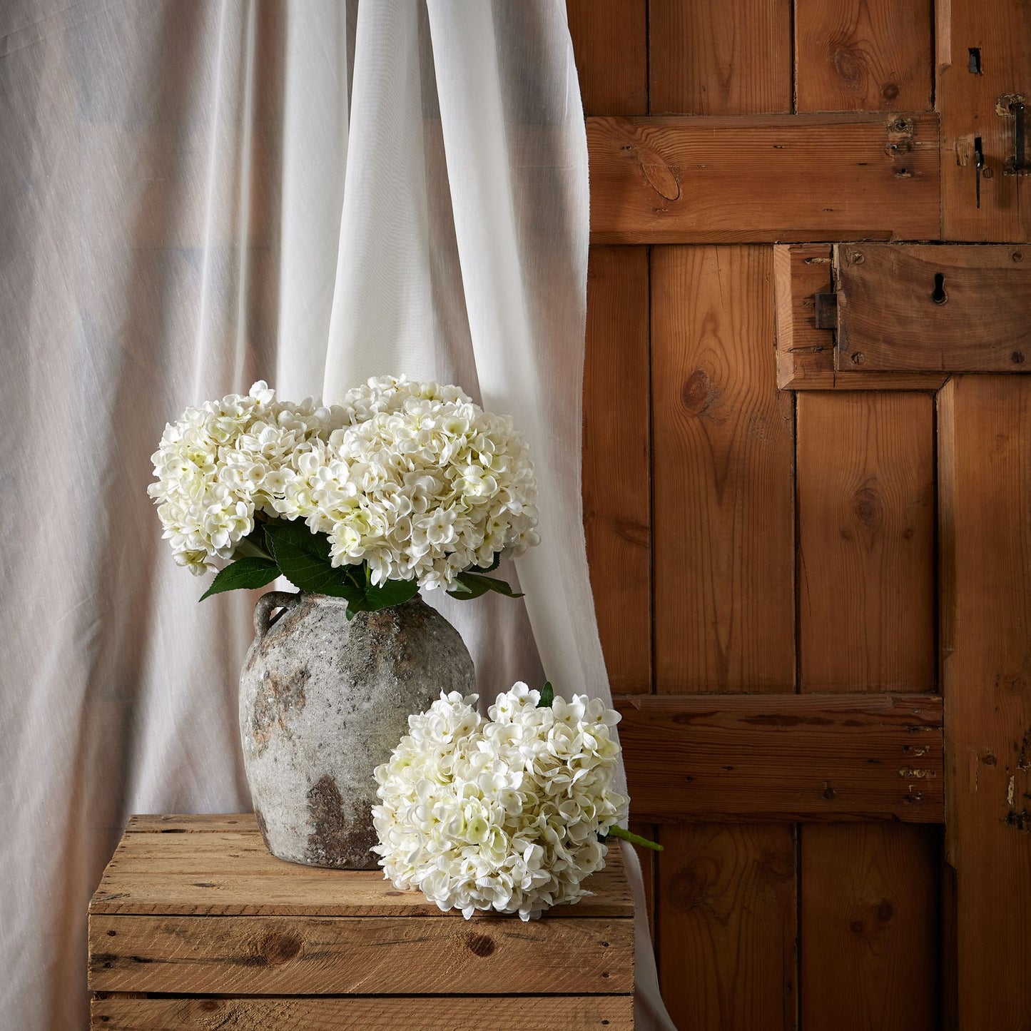 A bunch of pristine white artificial hydrangeas arranged in a textured gray vase, set against a rustic wooden backdrop and soft white drapery.