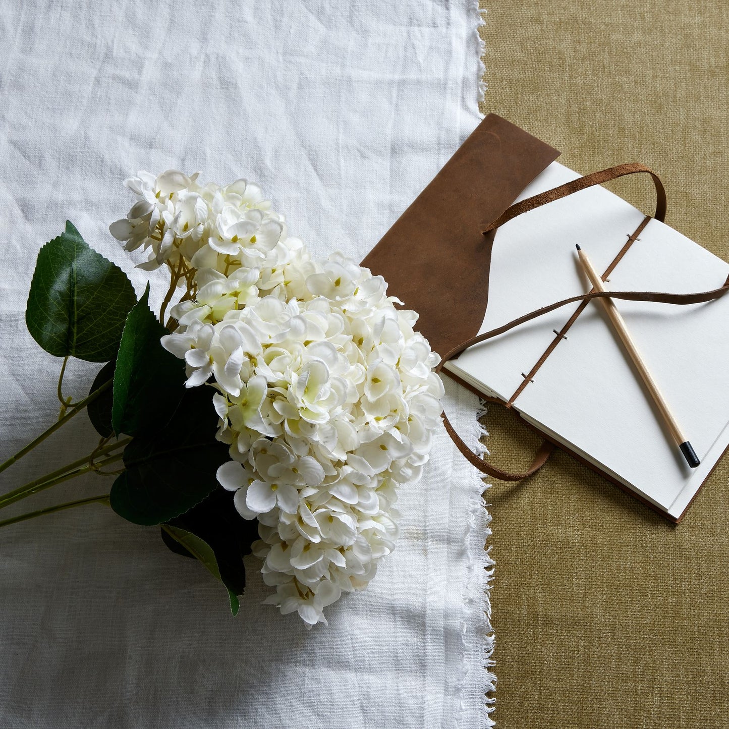 A lush bunch of white hydrangeas with detailed petals, accompanied by green leaves, resting on textured linens beside a leather notebook.