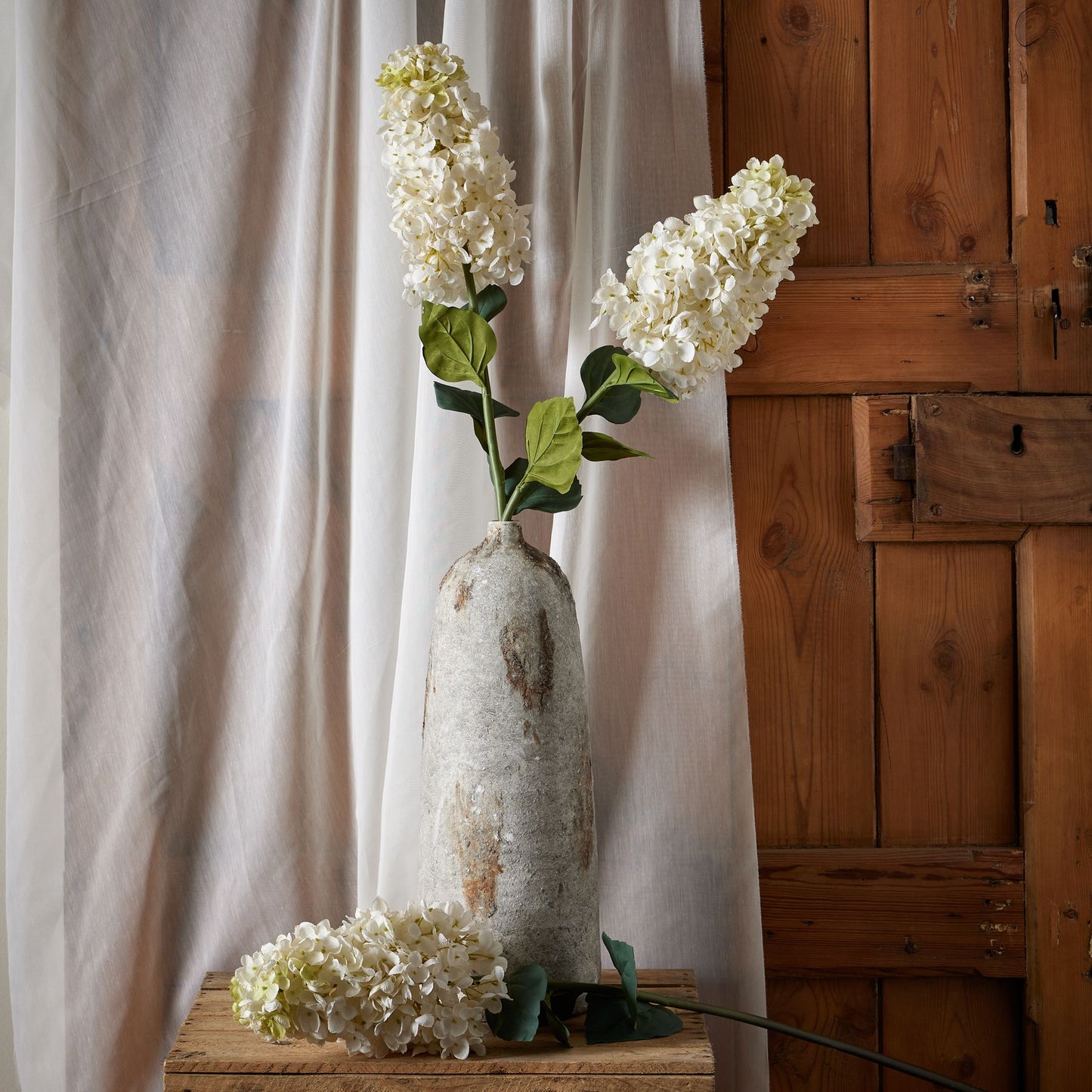 White spear hydrangea stems in a textured gray vase, accented by green leaves, enhance elegant home decor against a wooden backdrop.