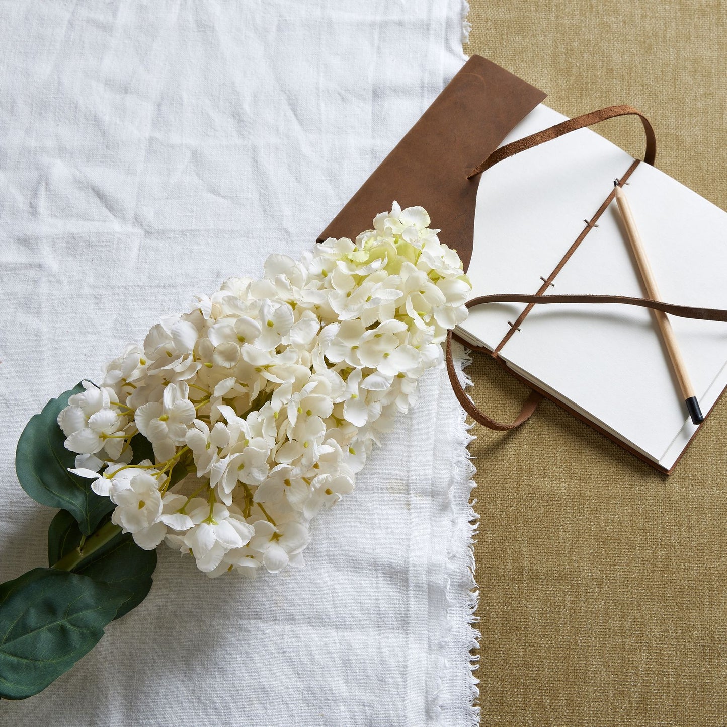 Elegant white spear hydrangea stem with lush green leaves, set on textured fabrics alongside a notebook and pen.
