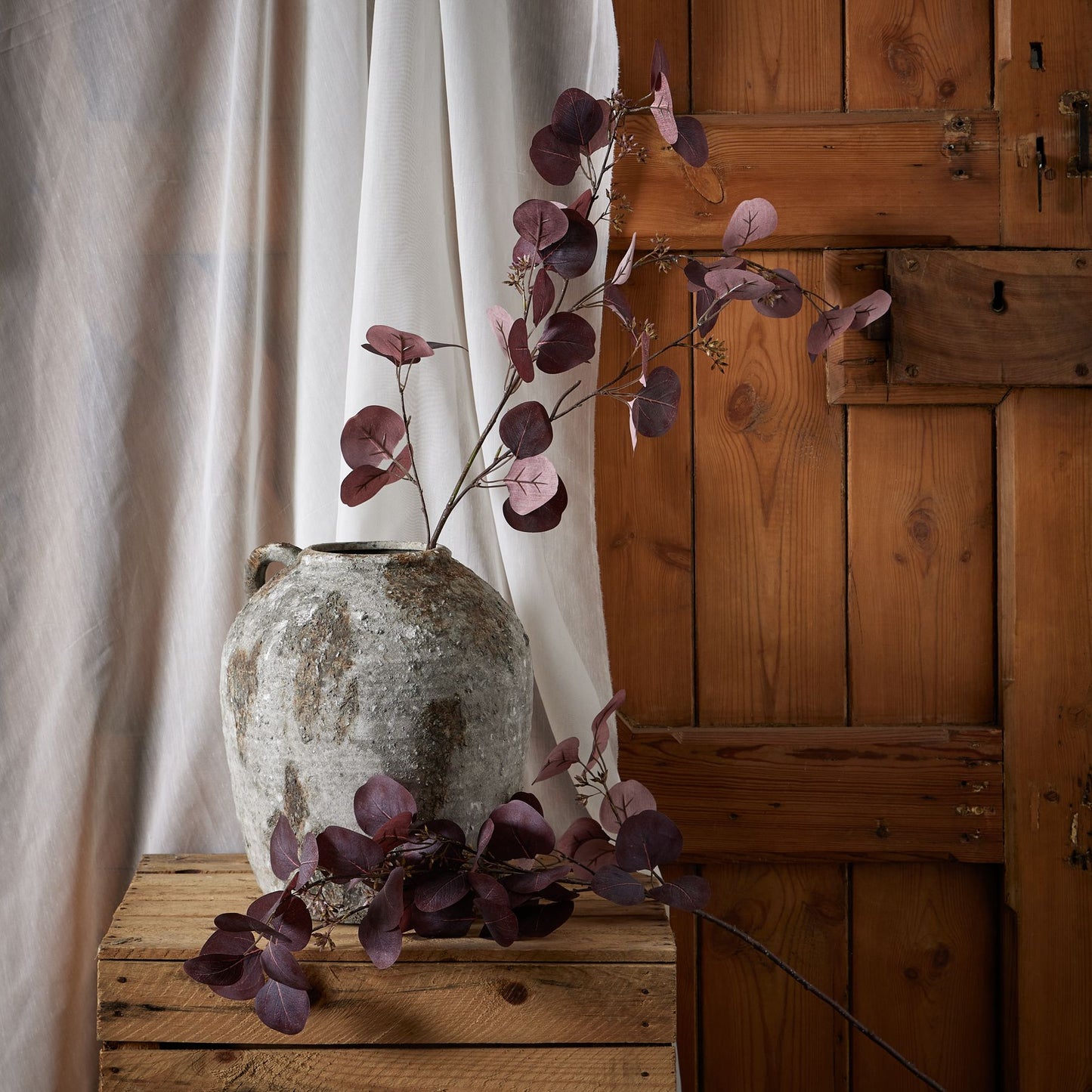Burgundy berried eucalyptus in an aged ceramic vase, set against warm wooden tones, adds autumn elegance and visual depth.
