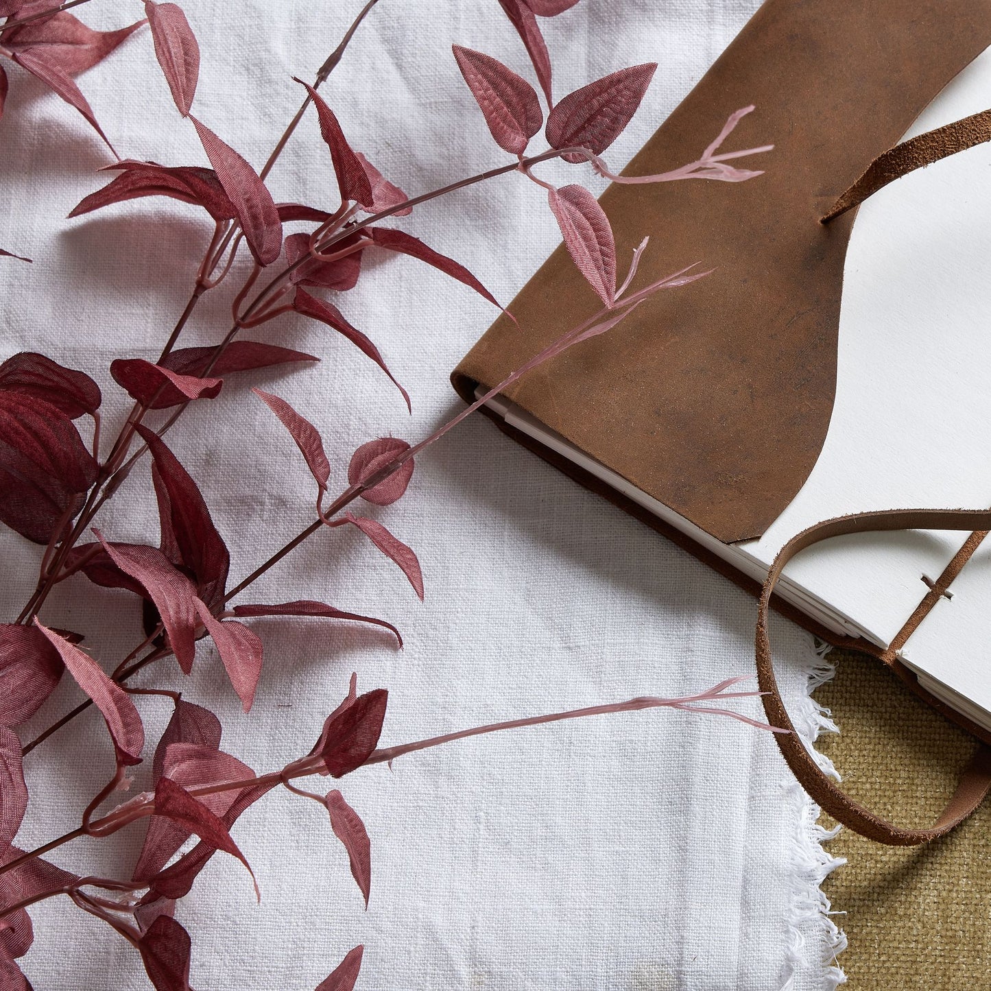Rich burgundy leaves gracefully arranged beside a brown leather notebook on a textured white and linen background.