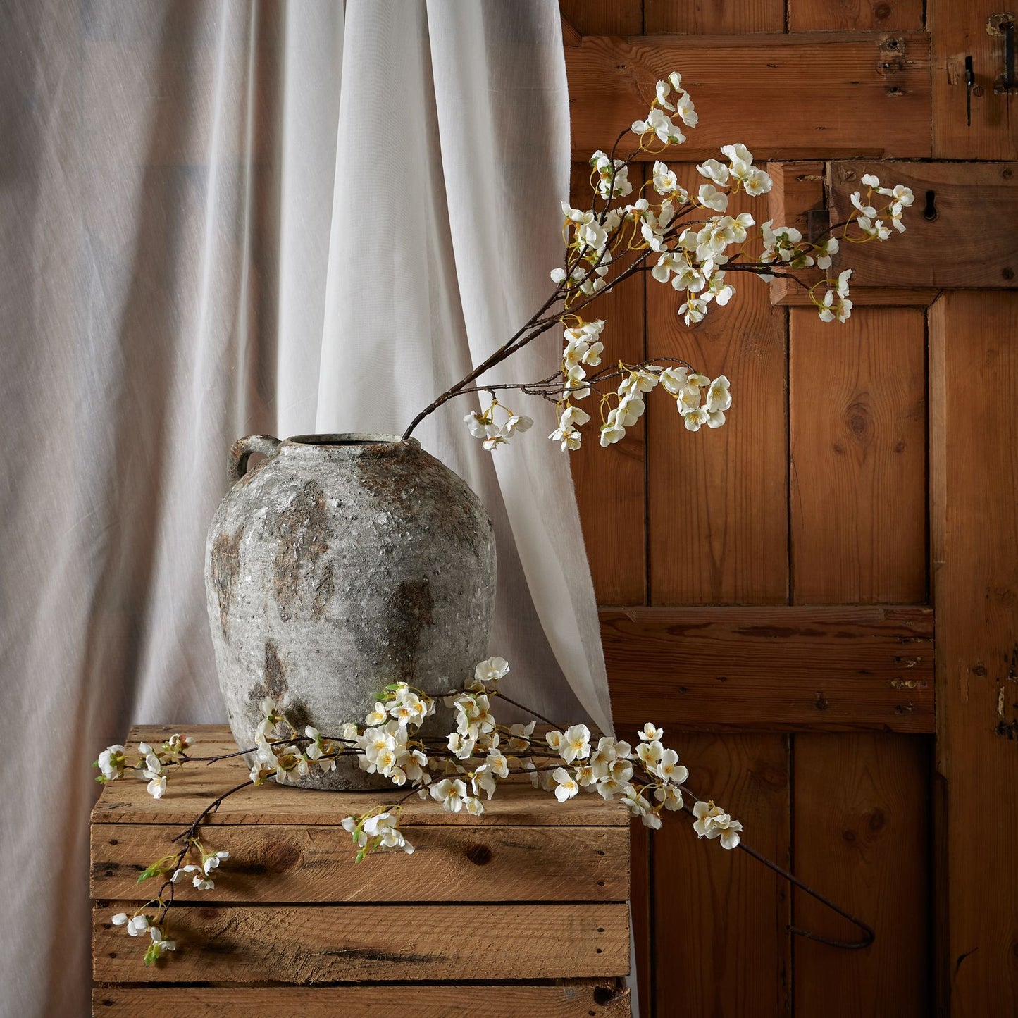 Delicate white Japanese blossoms on a slender branch, arranged in a textured gray vase atop a wooden crate, against a rustic backdrop.