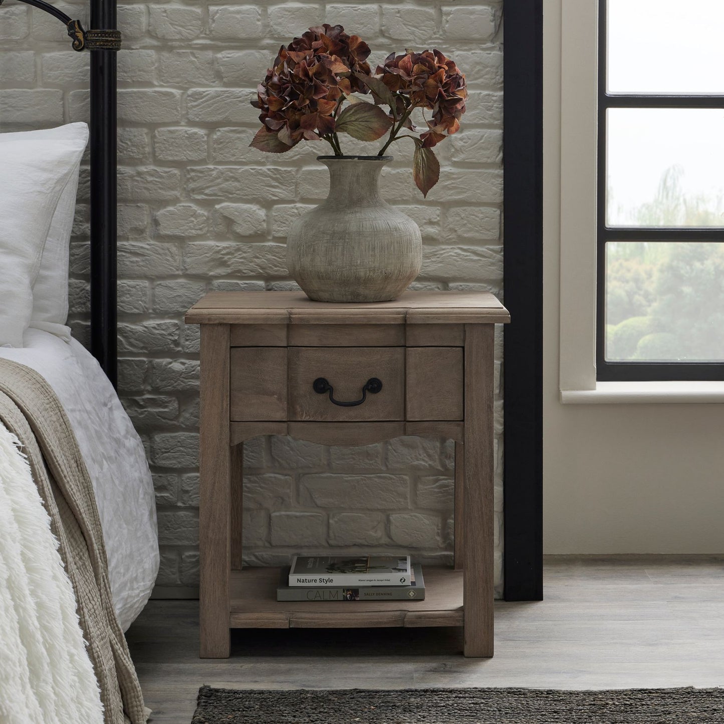 Rustic wooden side table with a washed finish, featuring an elegant drawer, shelf, and black iron handles, adorned with a vase of dried flowers.