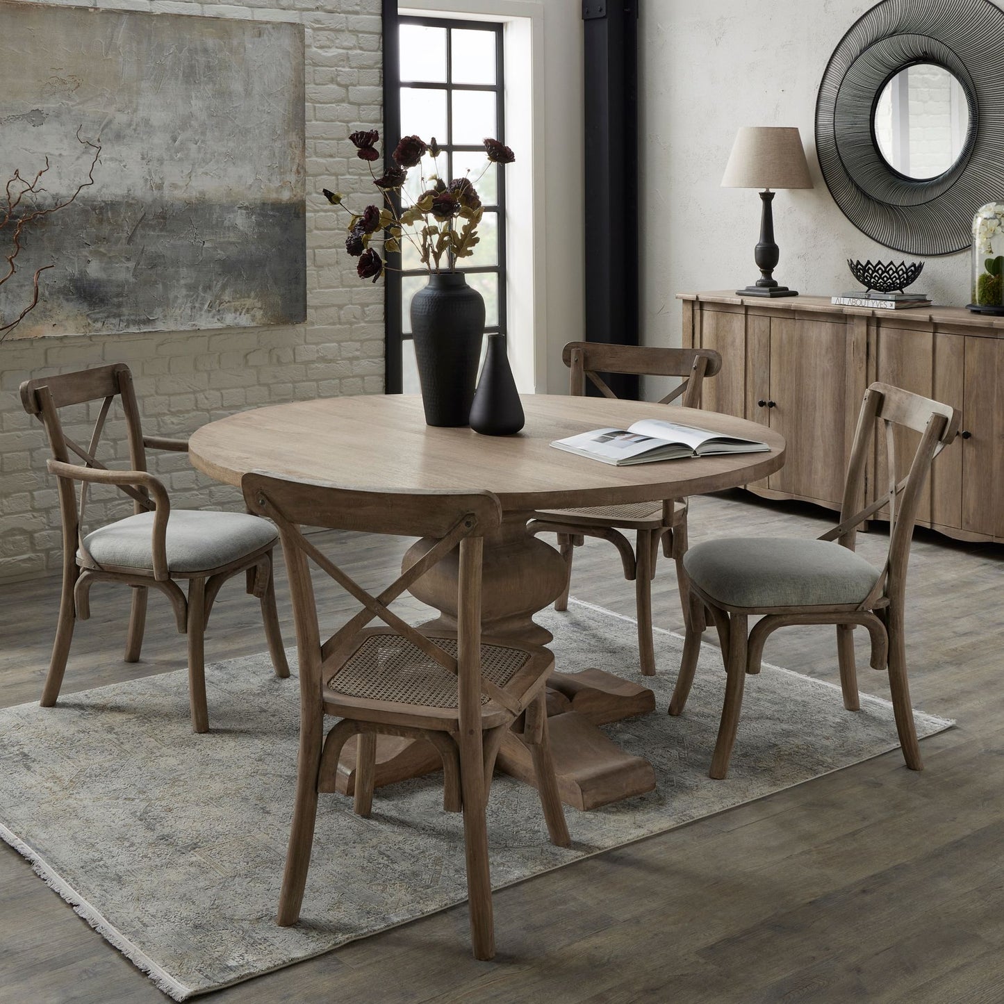 Round wooden pedestal dining table with a washed finish, complemented by light grey upholstered chairs, set in an elegant dining space.
