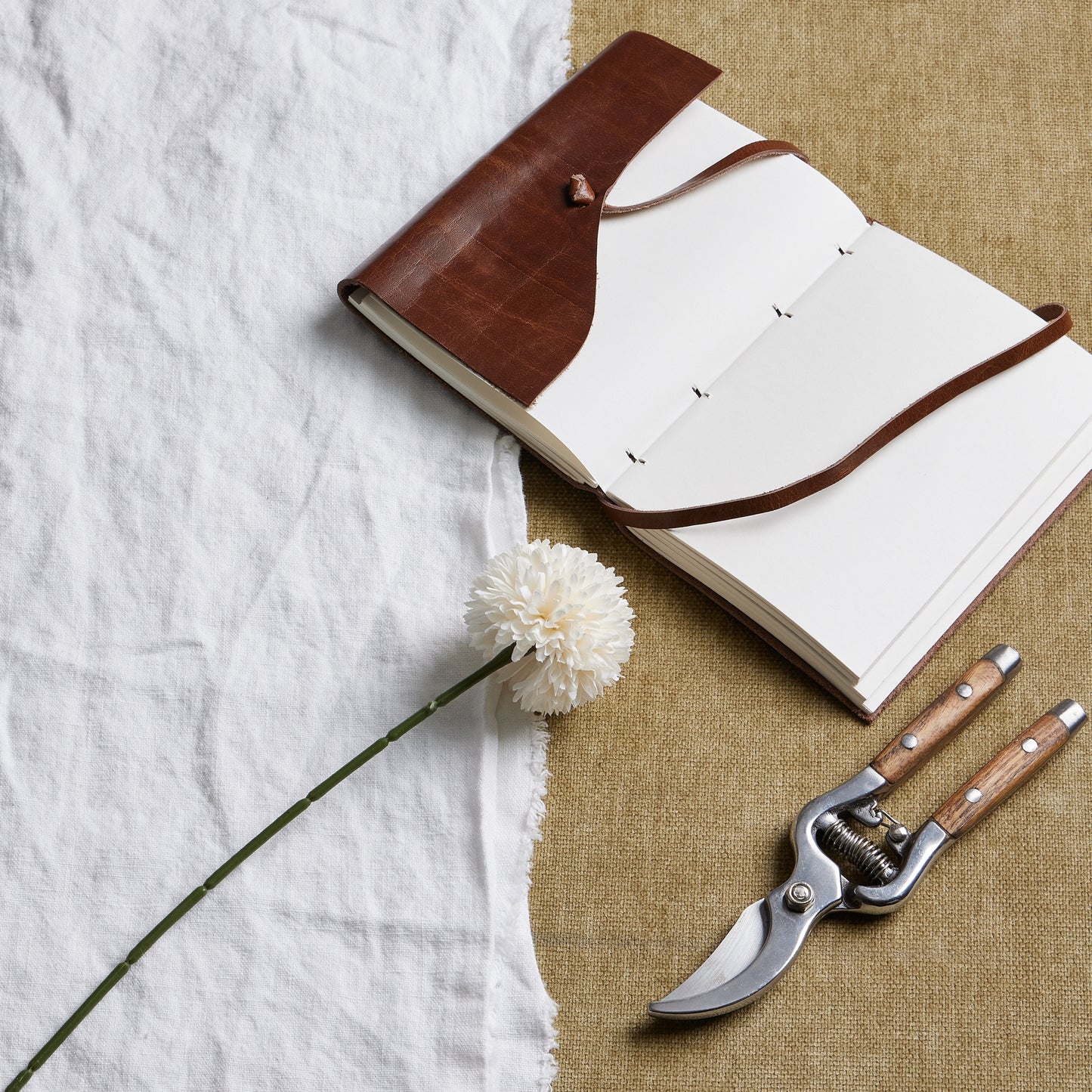 White short chrysanthemum with lifelike petals on a green stem, styled alongside a leather notebook and garden shears on textured fabric.