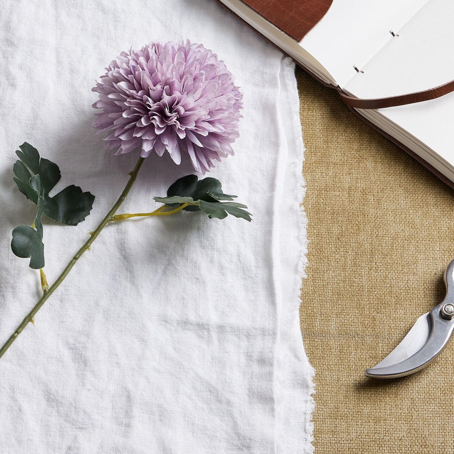 Light purple artificial chrysanthemum with layered petals and green leaves, resting on a textured beige surface.