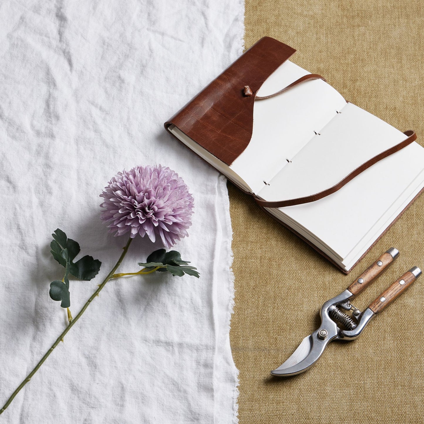 Light purple chrysanthemum with layered petals beside a leather notebook and garden shears on a textured surface.