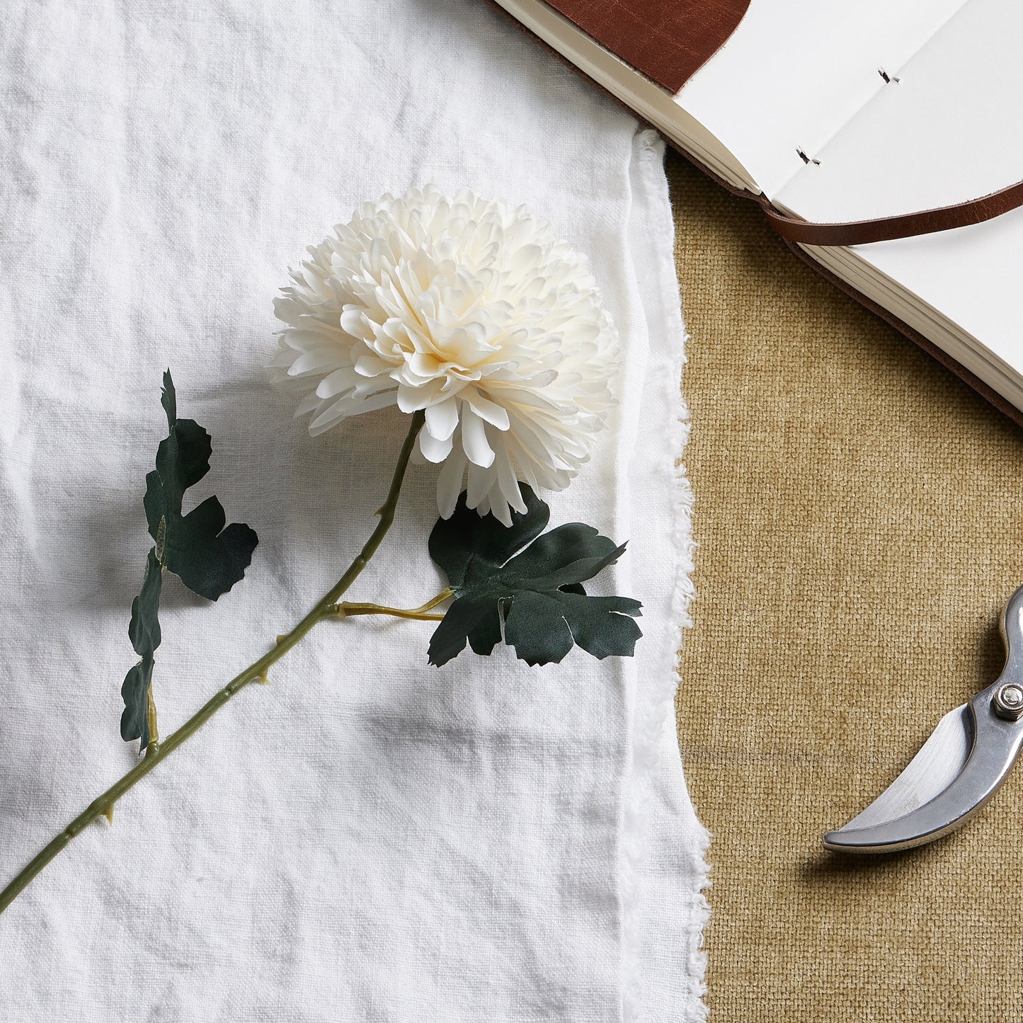 Lifelike white chrysanthemum with delicate petals and green foliage, displayed on a textured surface with a journal and scissors.