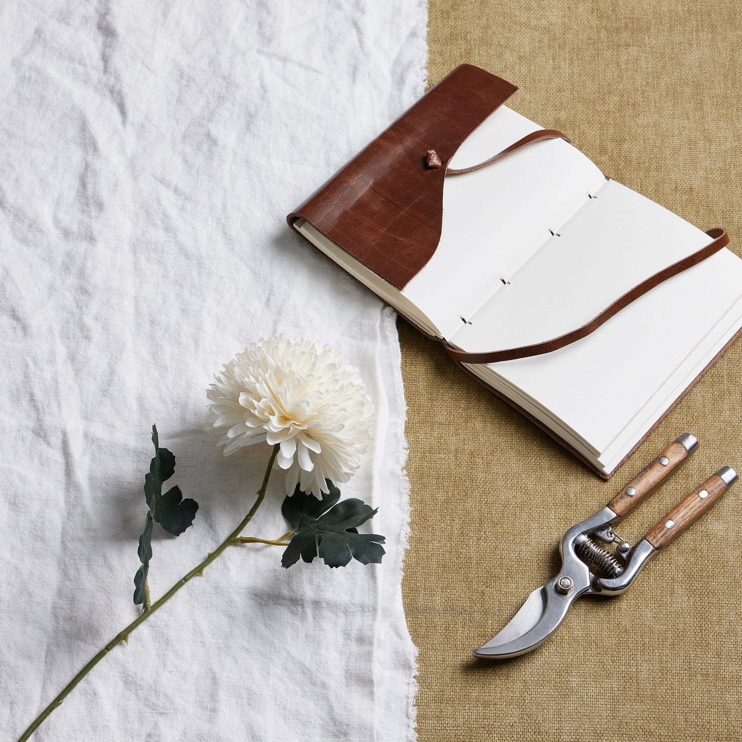 Lifelike white chrysanthemum on a textured fabric background, beside a brown leather notebook and gardening shears.