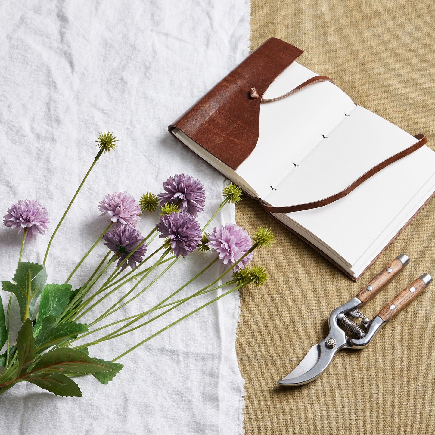 A bouquet of artificial purple chrysanthemums with layered petals lies next to a leather-bound notebook and gardening shears on a textured surface.