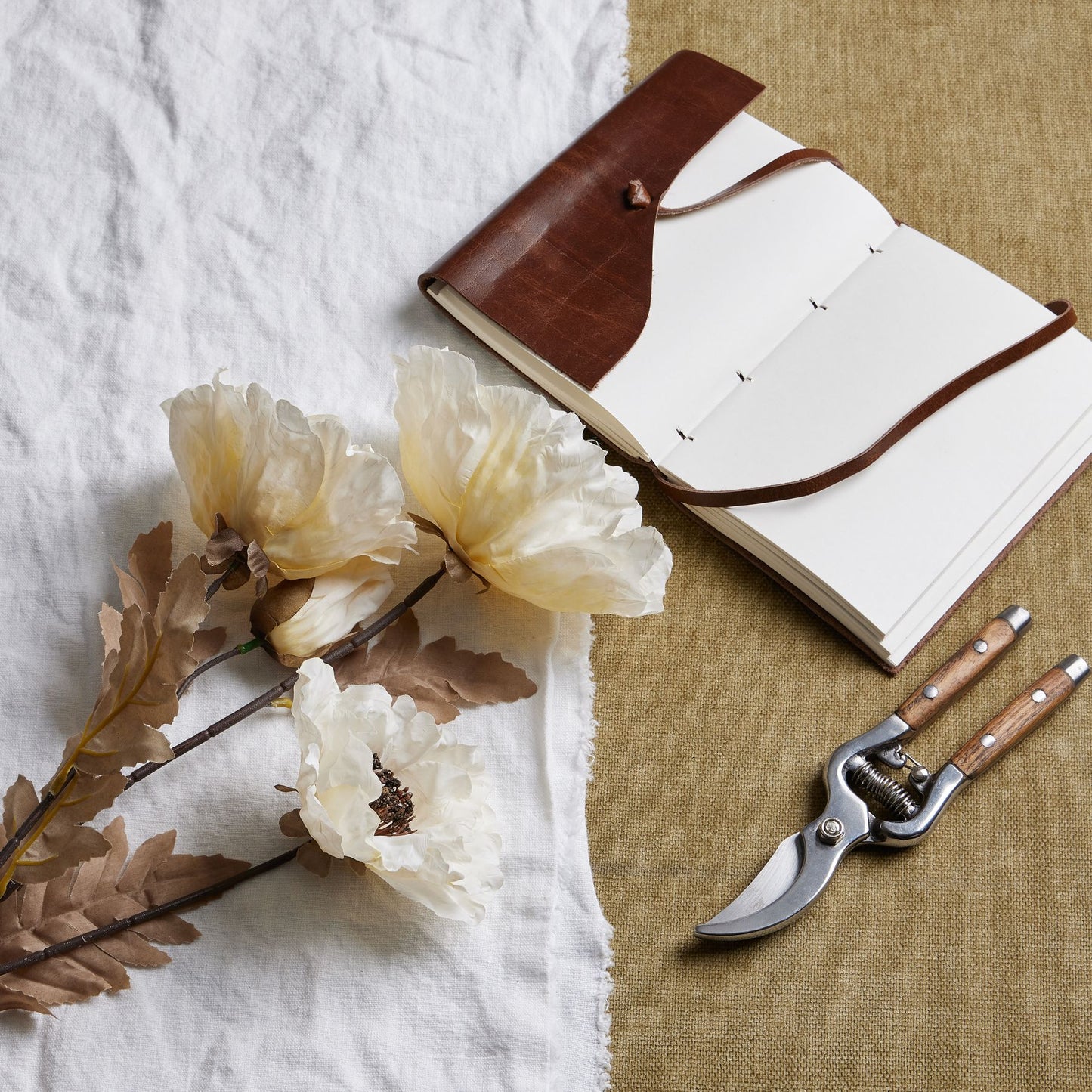 Cream-colored large poppy blooms with subtle brown accents, resting beside a brown notebook and pruning shears on a textured background.