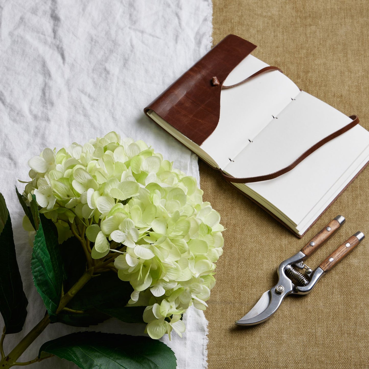 Oversized green hydrangea with hand-painted petals, nestled beside a leather notebook and pruning shears on textured fabric.
